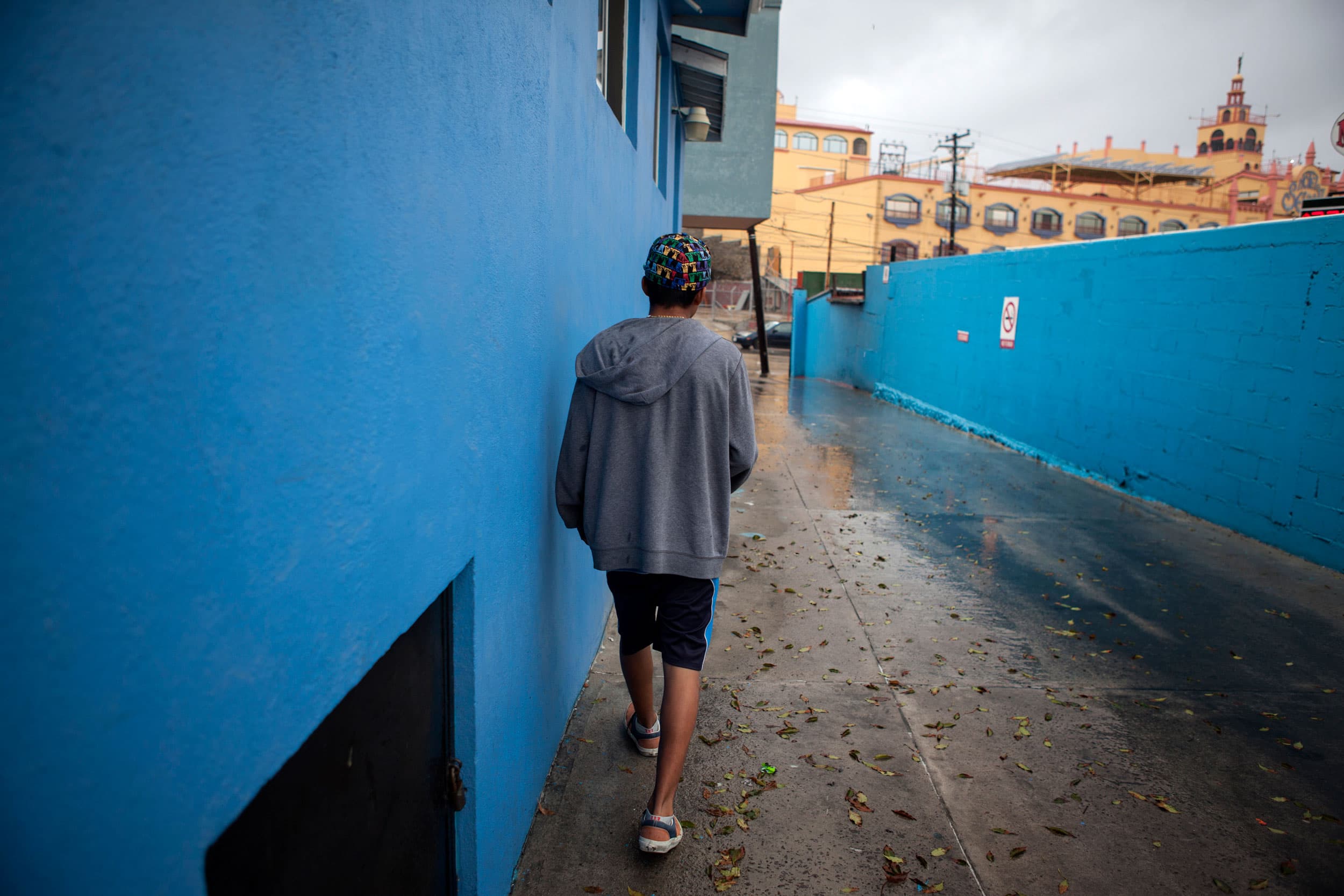 A teenage boy walks down a driveway past a bright blue building. His back is to the camera.