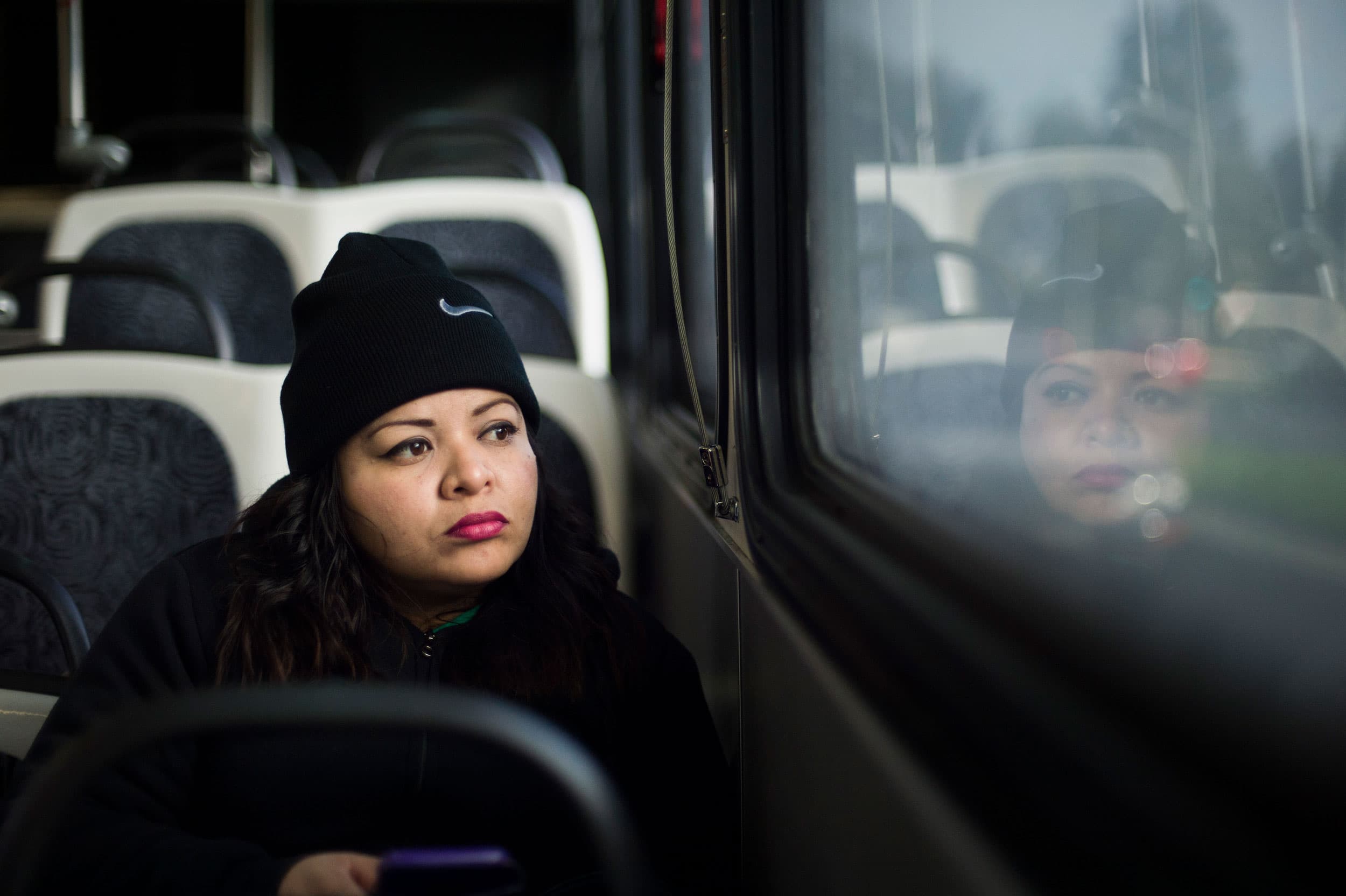 A woman sits on a city bus, looking out the window. Her face is reflected back in the glass.
