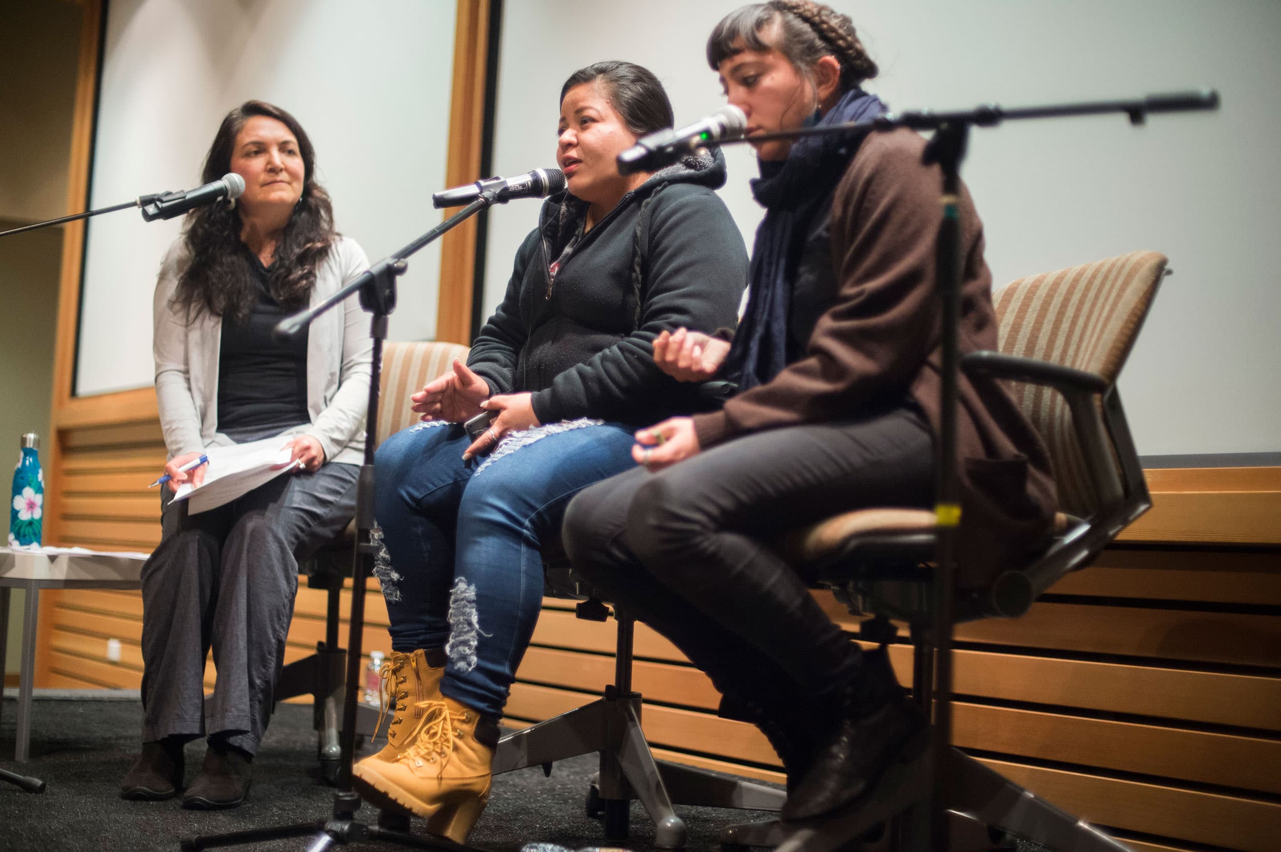Three women sit on chairs on a stage, each in front of a small microphone