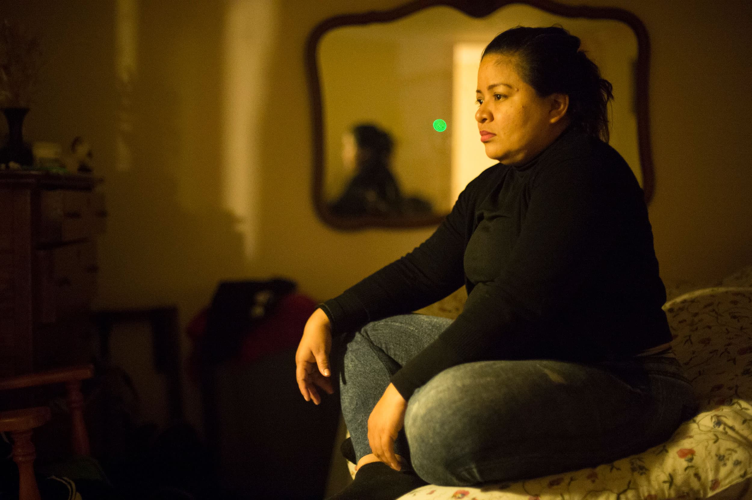 A woman sits on a bed in a dark room. She is lit by the streetlights flooding through a window.