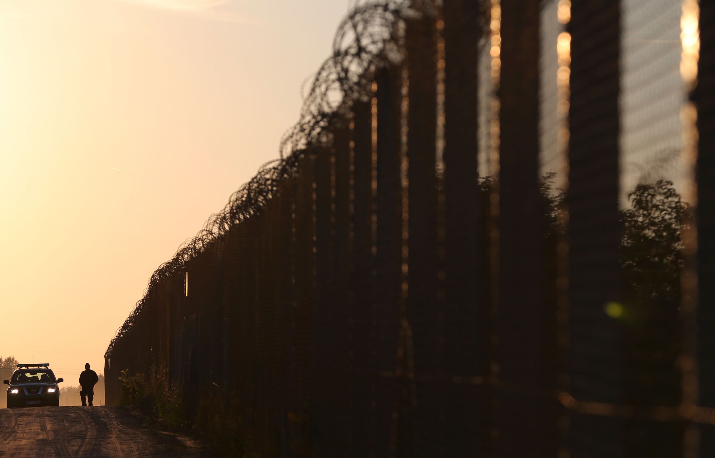 A police officer and a tall fence topped with razor wire is silhouetted against an orange sky.
