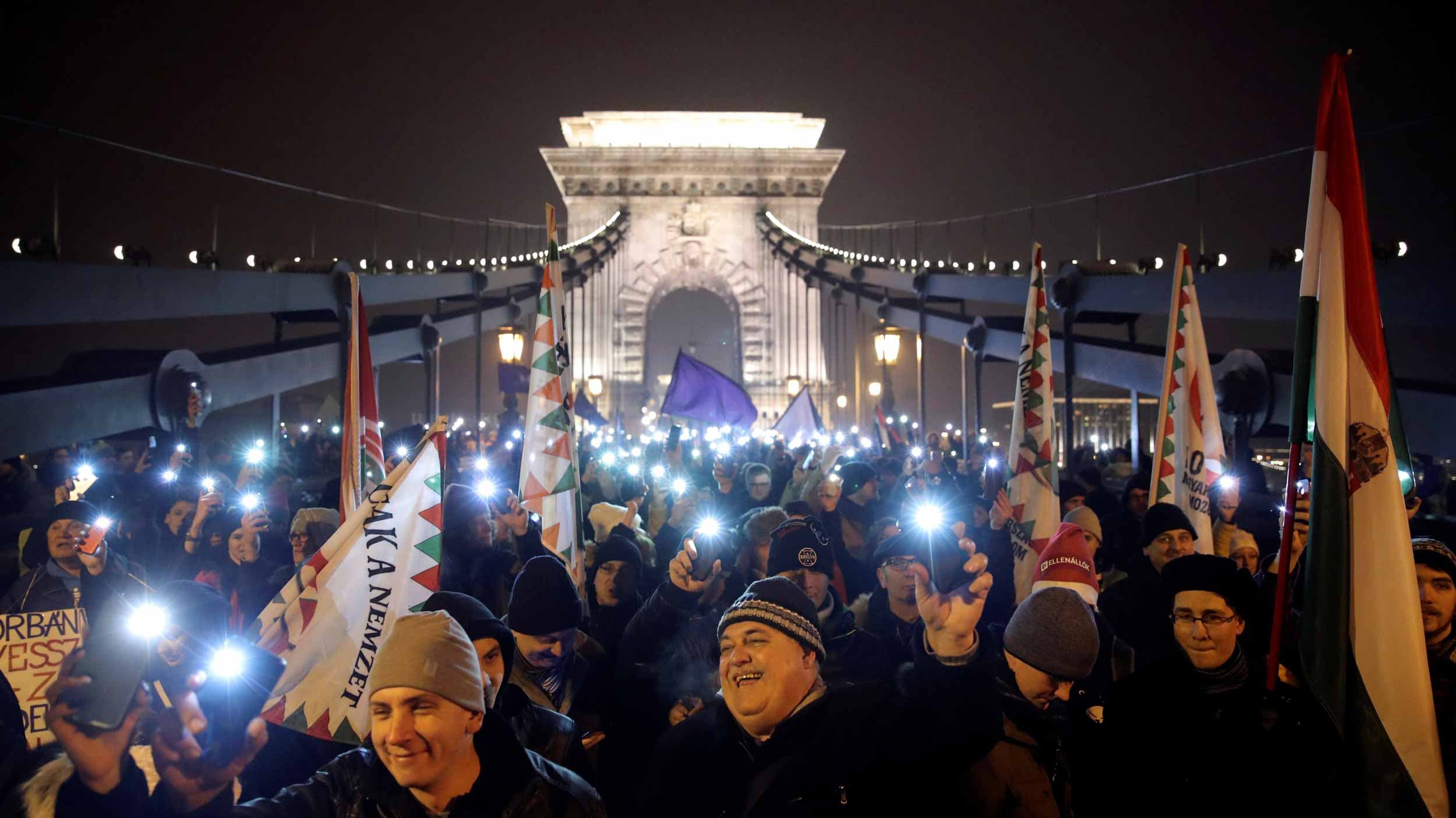 Protestors are crowded onto a bridge that is lit up behind them as they hold their cell phones in the air with the camera flashes turned on.