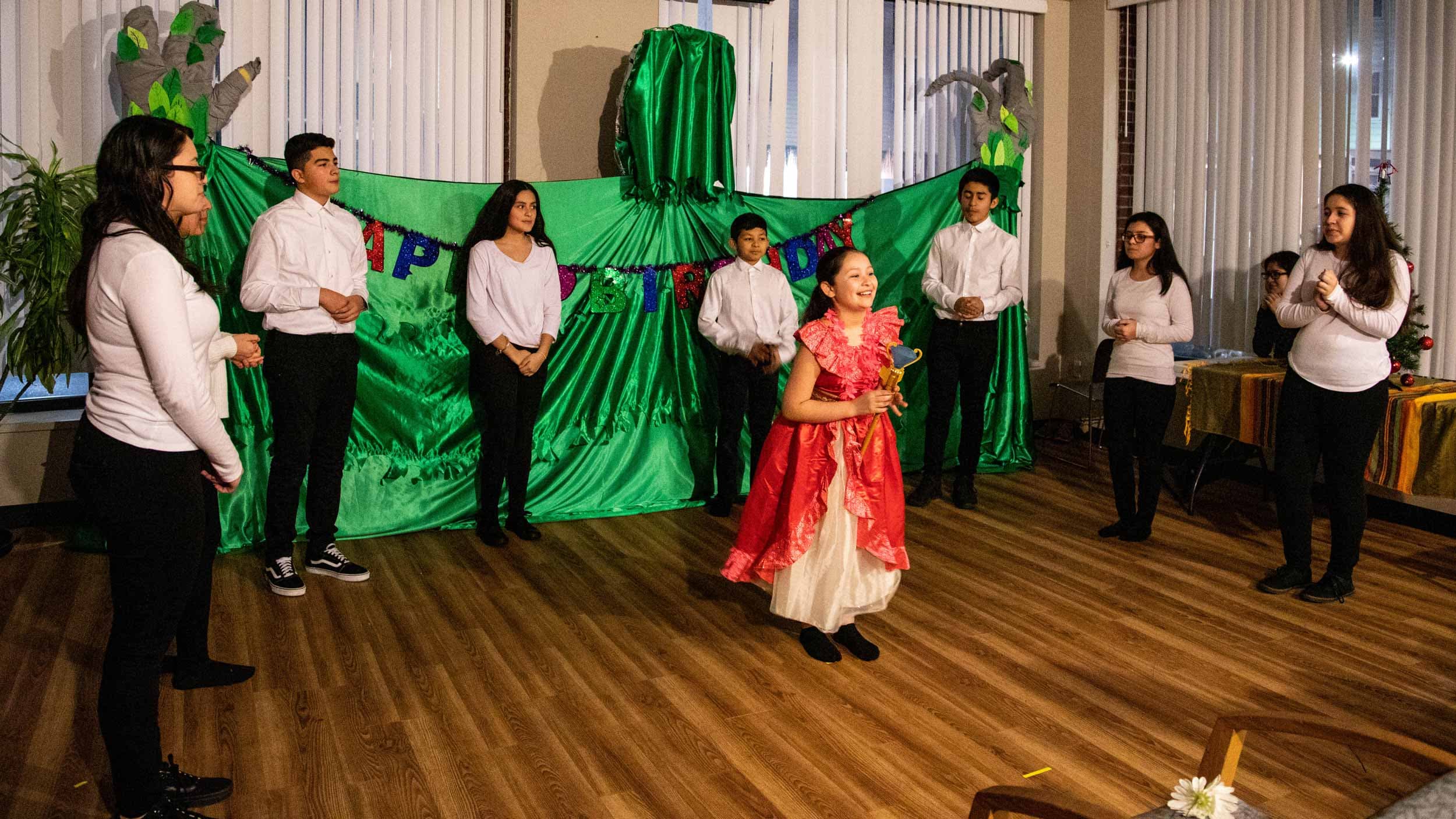 A girl wearing a red dress stands at the front of a room acting in a play