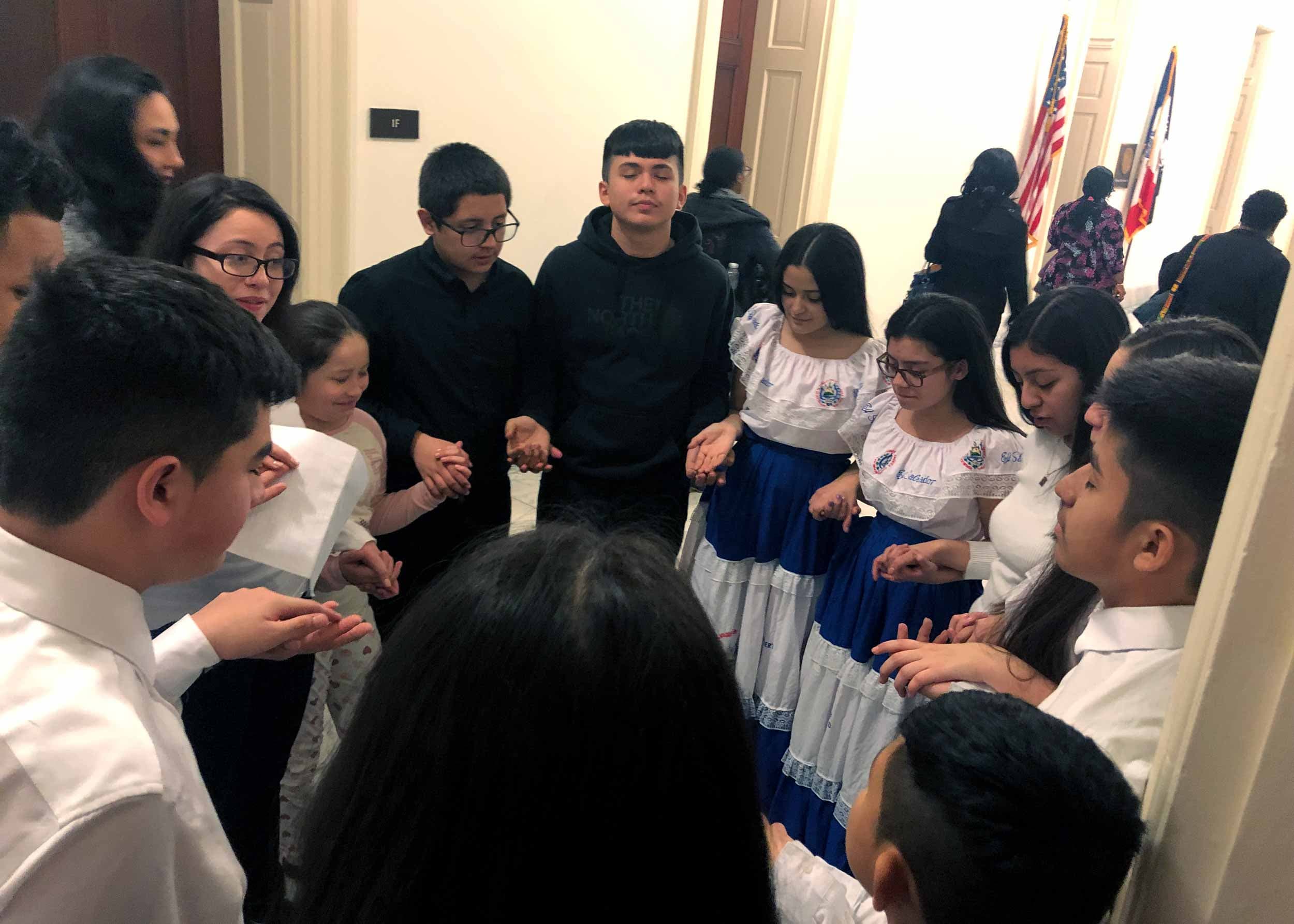 A group of children hold hands and bow their heads in prayer in an office hallway