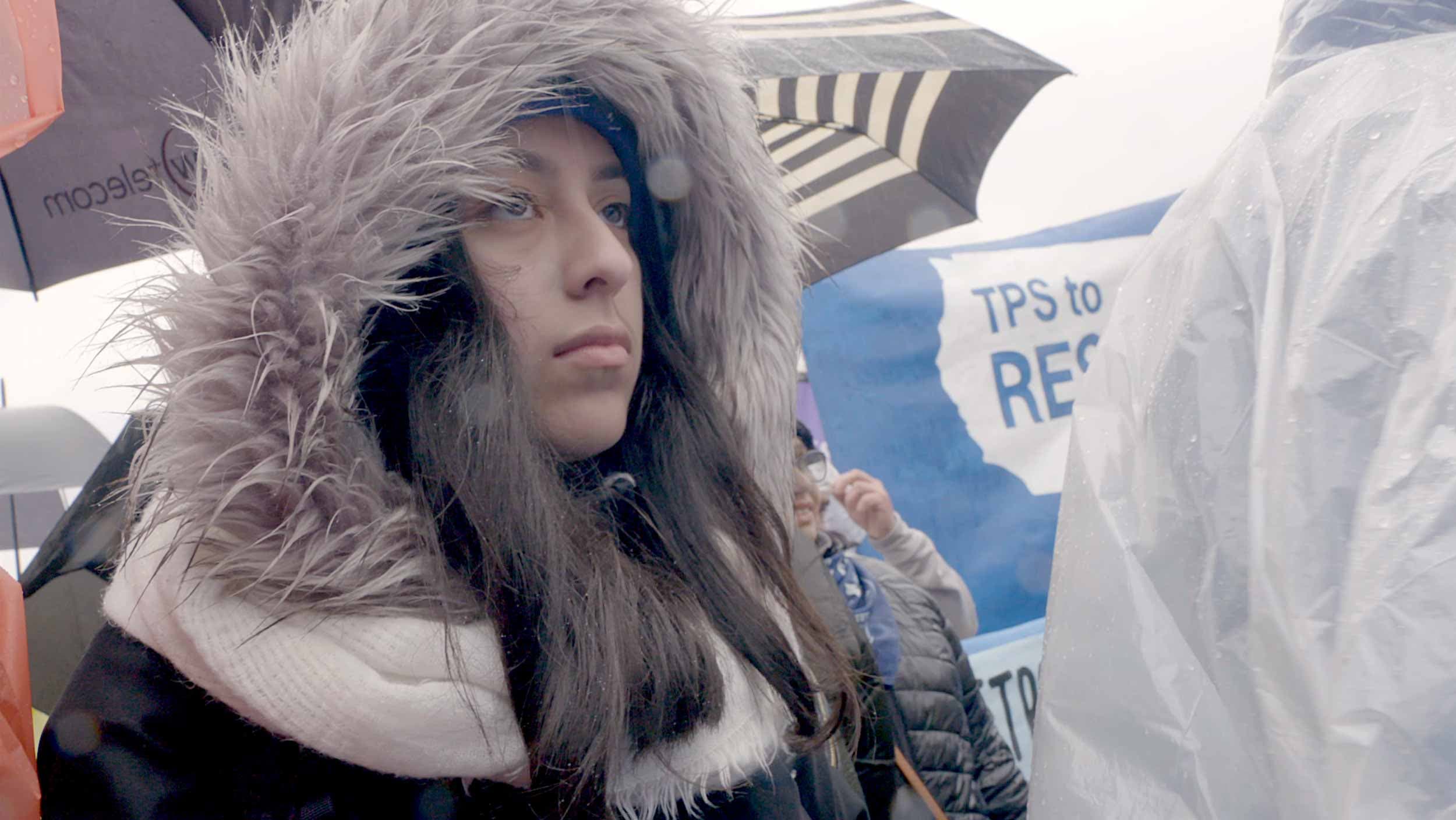 A close-up photo of a young woman wearing a furry hood at a TPS protest with blue signs in the background.