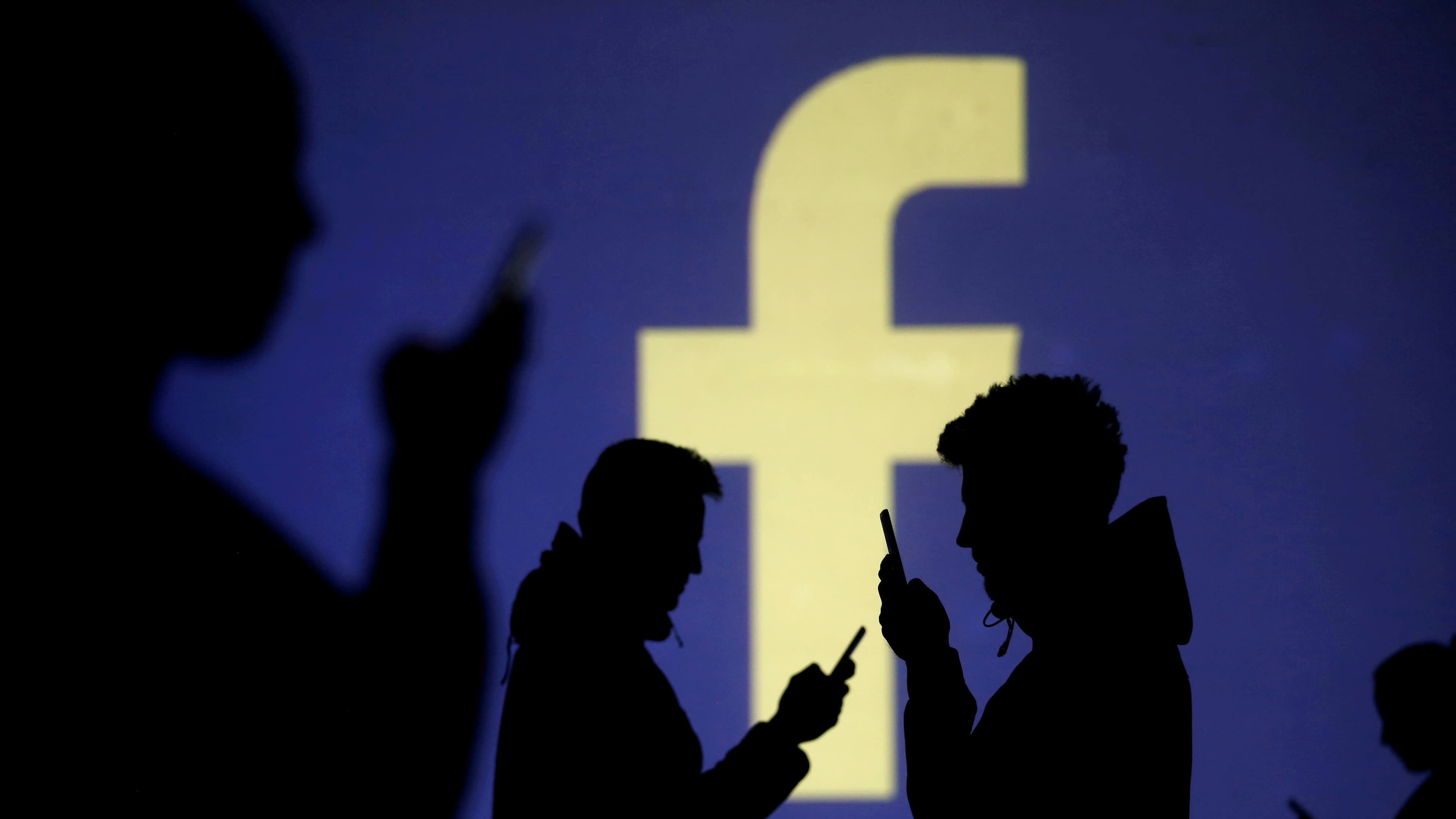 Silhouettes of mobile users are seen next to a screen projection of Facebook logo