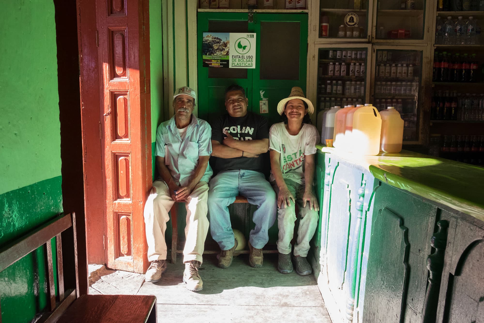Three men are shown sitting on a bench in a store with bottles on the wall.
