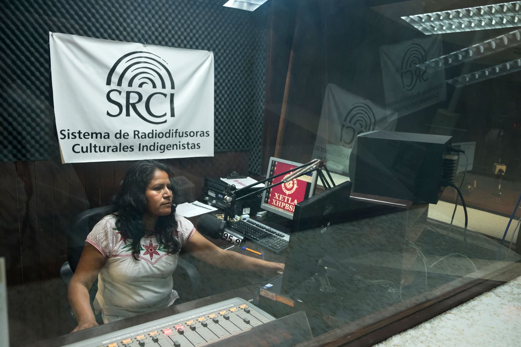 Eva Hernández is shown sitting behind clear class at a radio DJ board with a microphone.