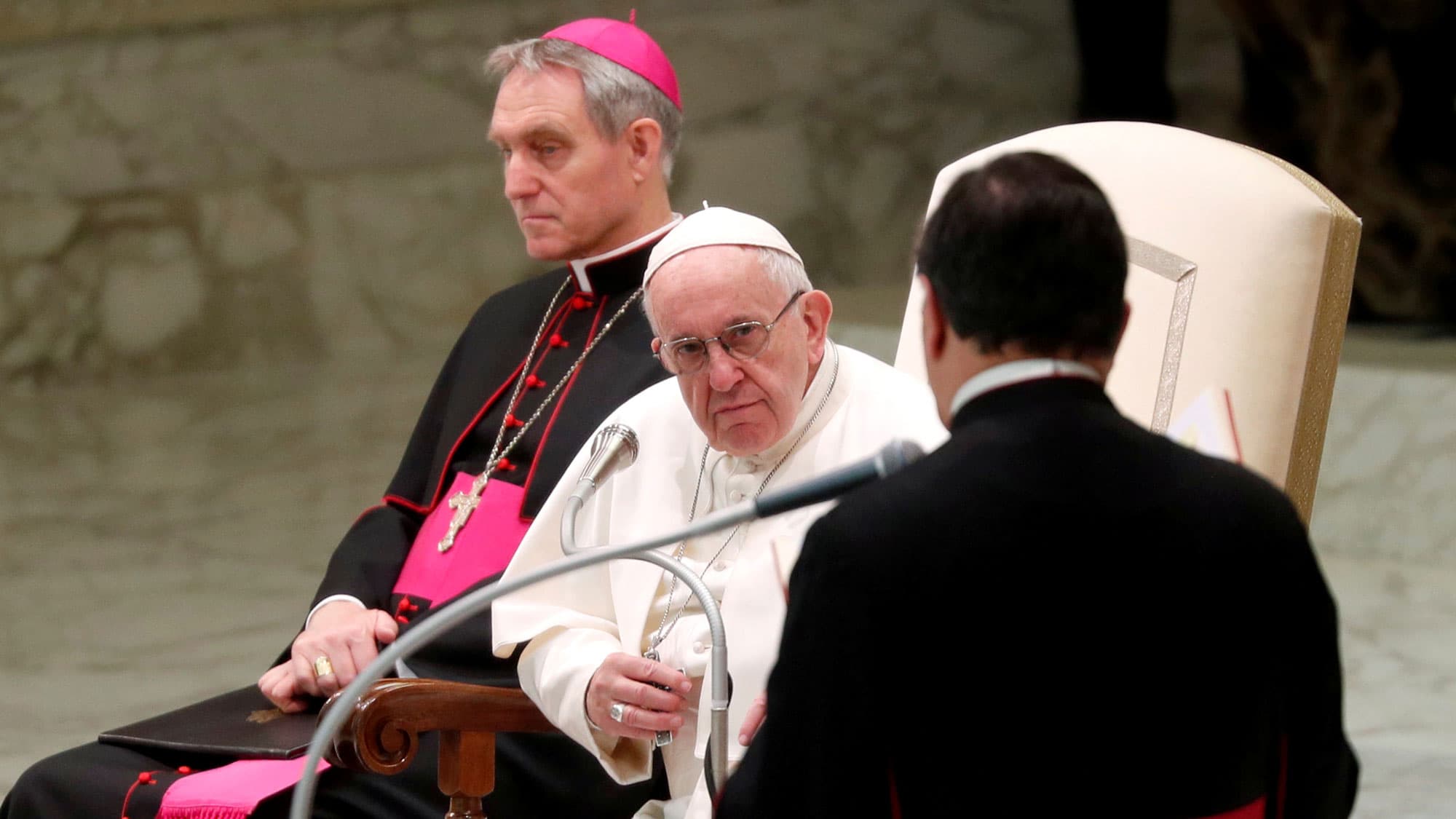 Pope Francis is shown wearing white while sitting behind a microphone.
