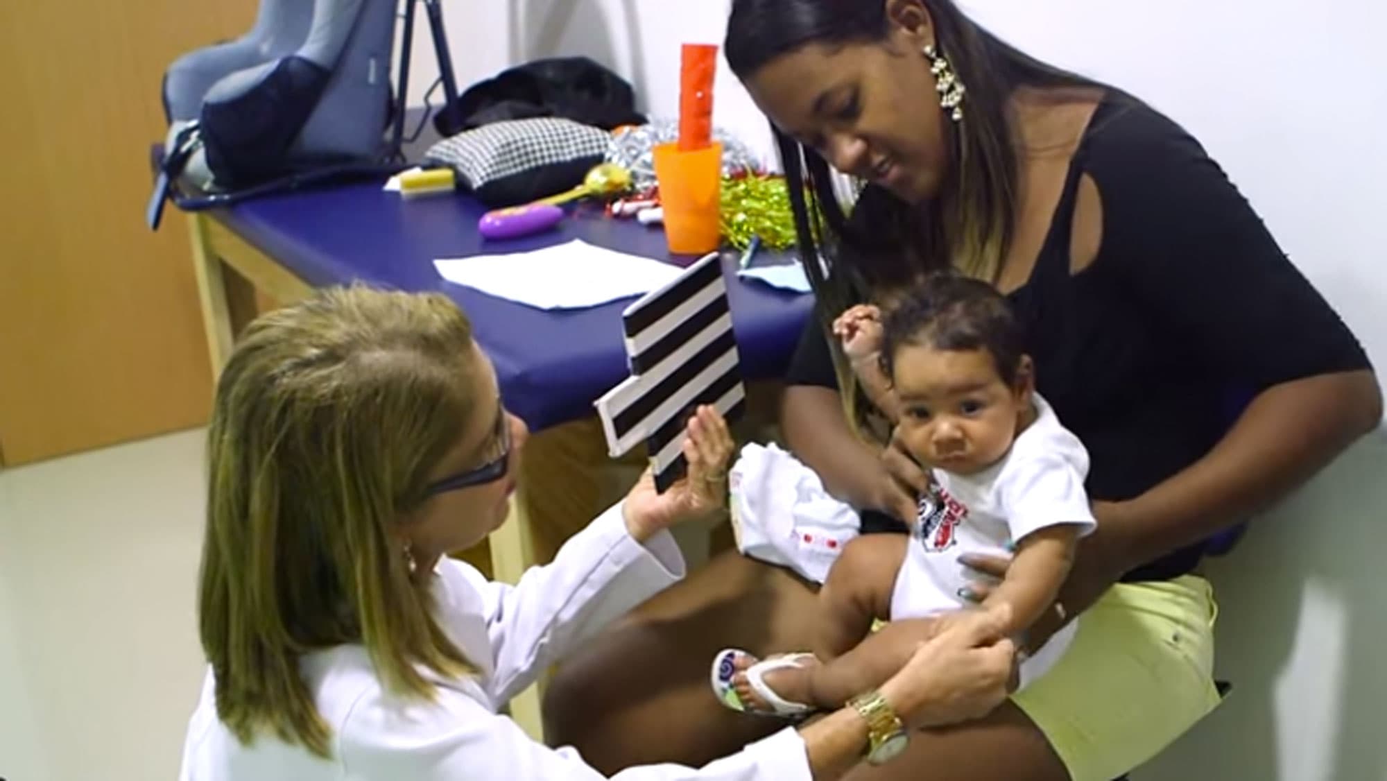 Germana Soares is shown holding her child next to a doctor holding a black and white striped card.