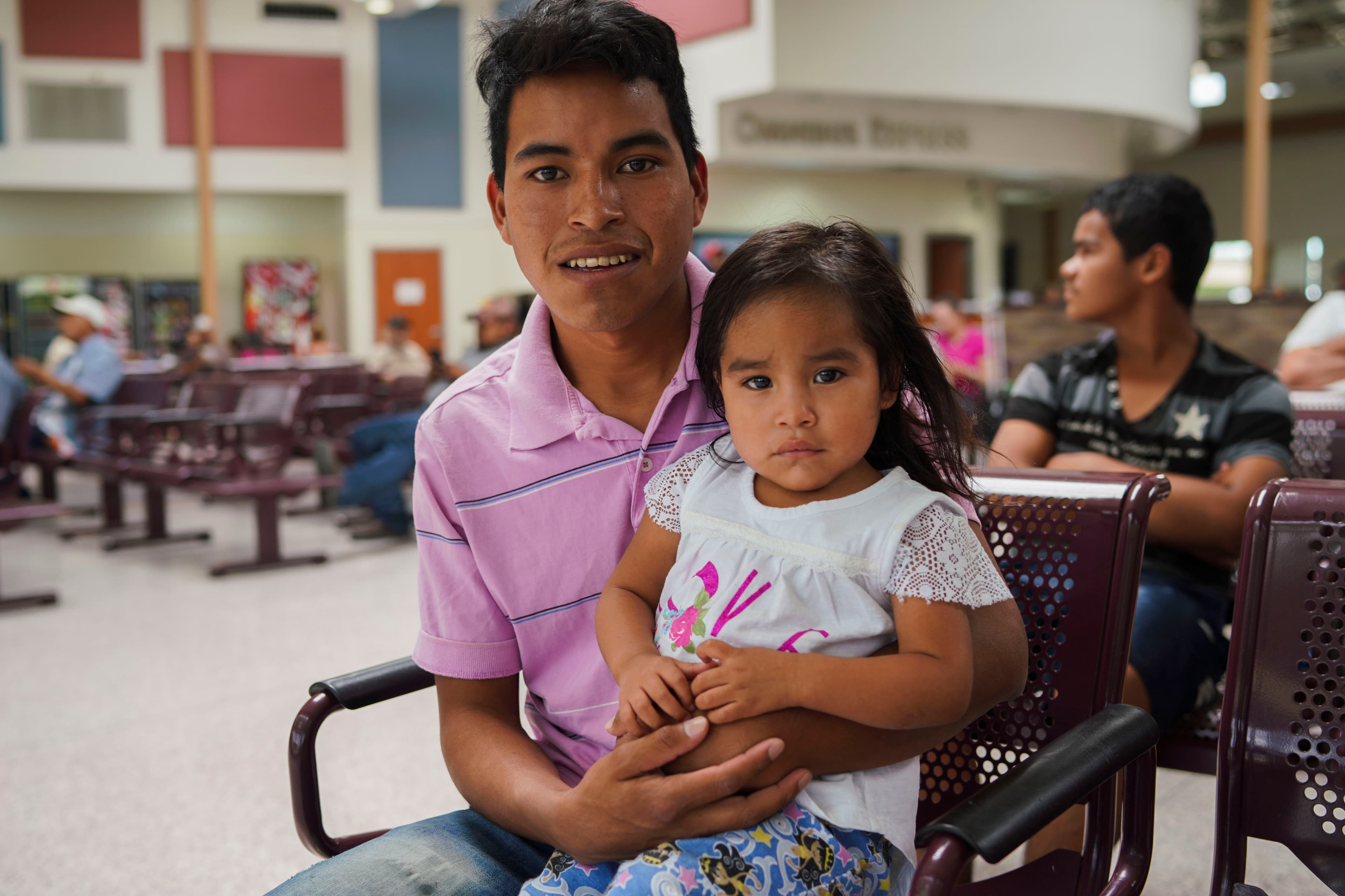 A portrait of a father wearing a pink shirt and his infant daughter who is wearing white.
