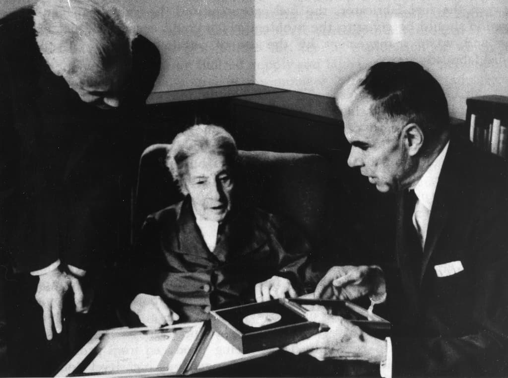 An elderly white woman sits between two white men presenting an award.