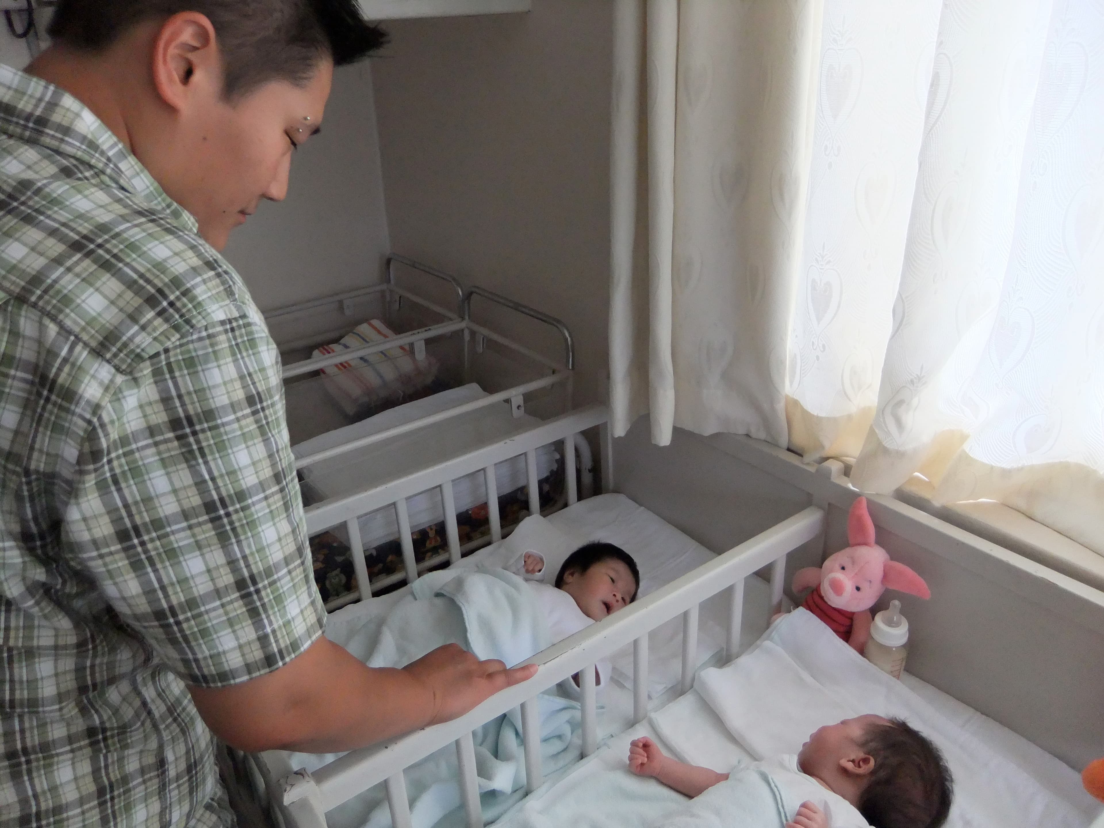A woman stands over two cribs with babies in them.