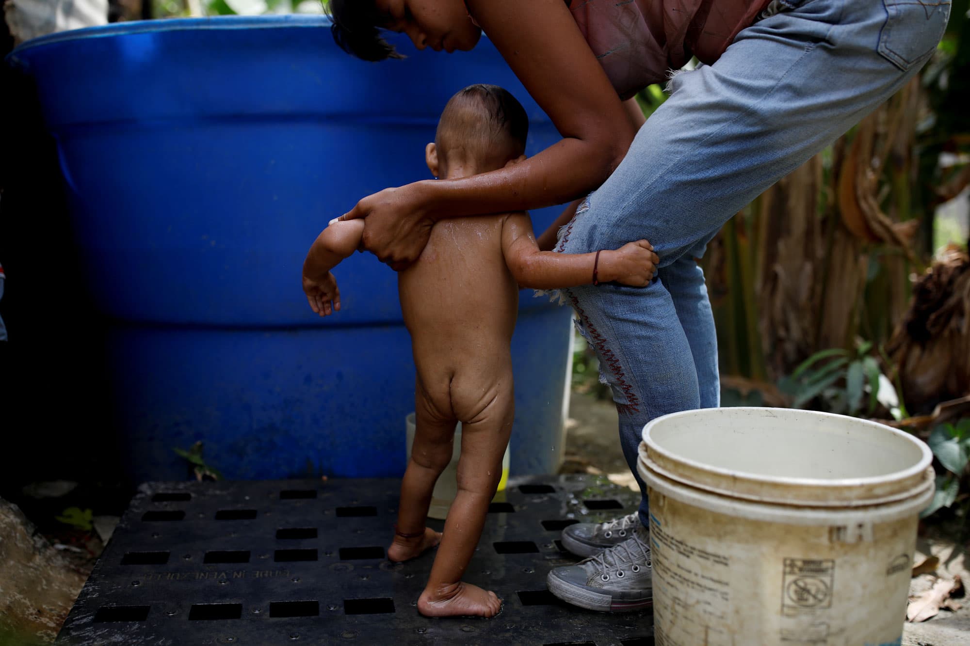 A woman is shown holding the hands of her naked baby during a bath.