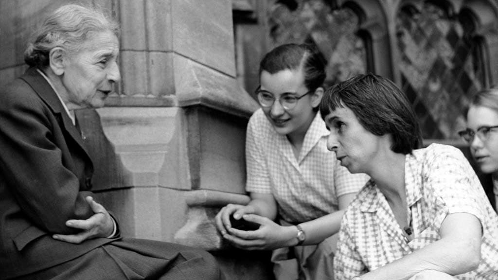 An elderly white woman sits on a step, talking with two younger white women.