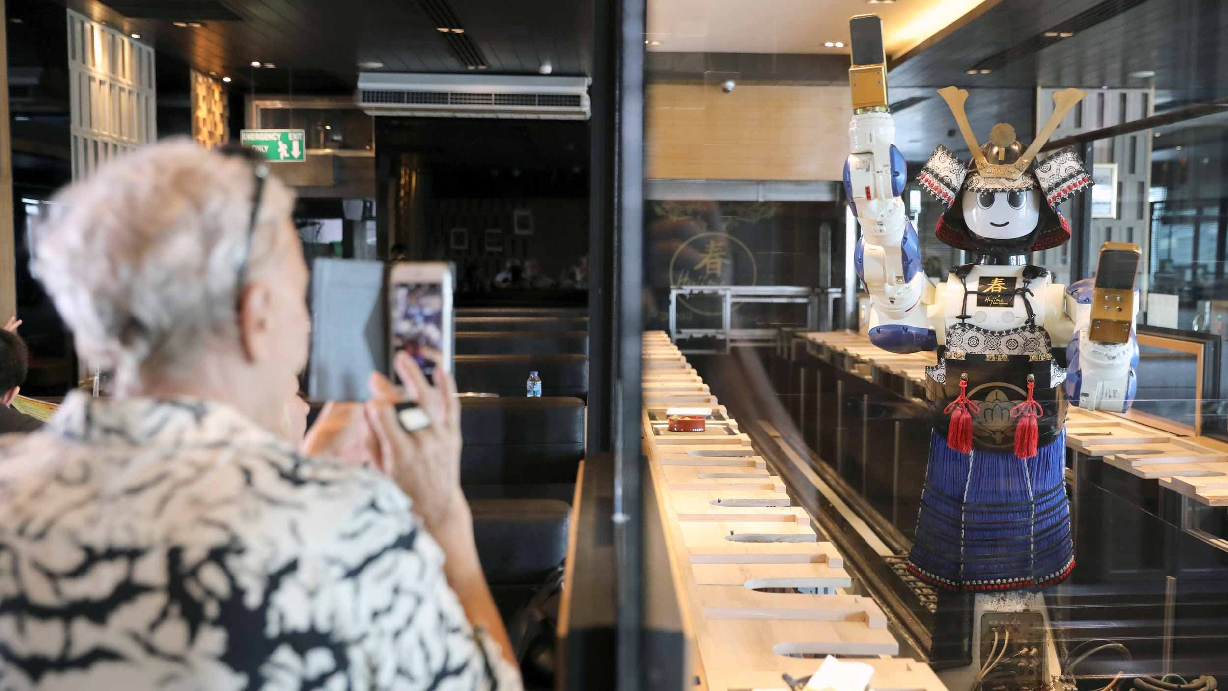 A man, left, takes a photo of a colorful robot that is also working as waitstaff in a restaurant.