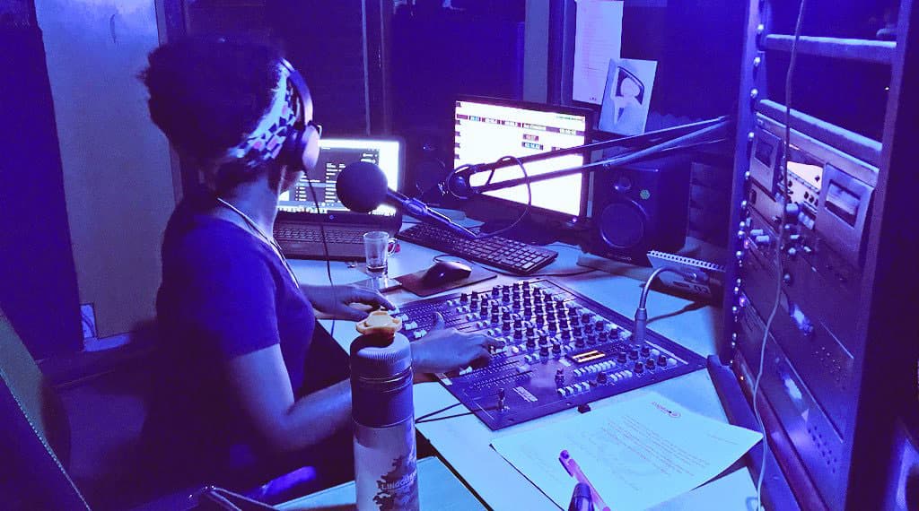 A woman with her back to the camera wears headphones and talks into a microphone while sliding up switches on a control board