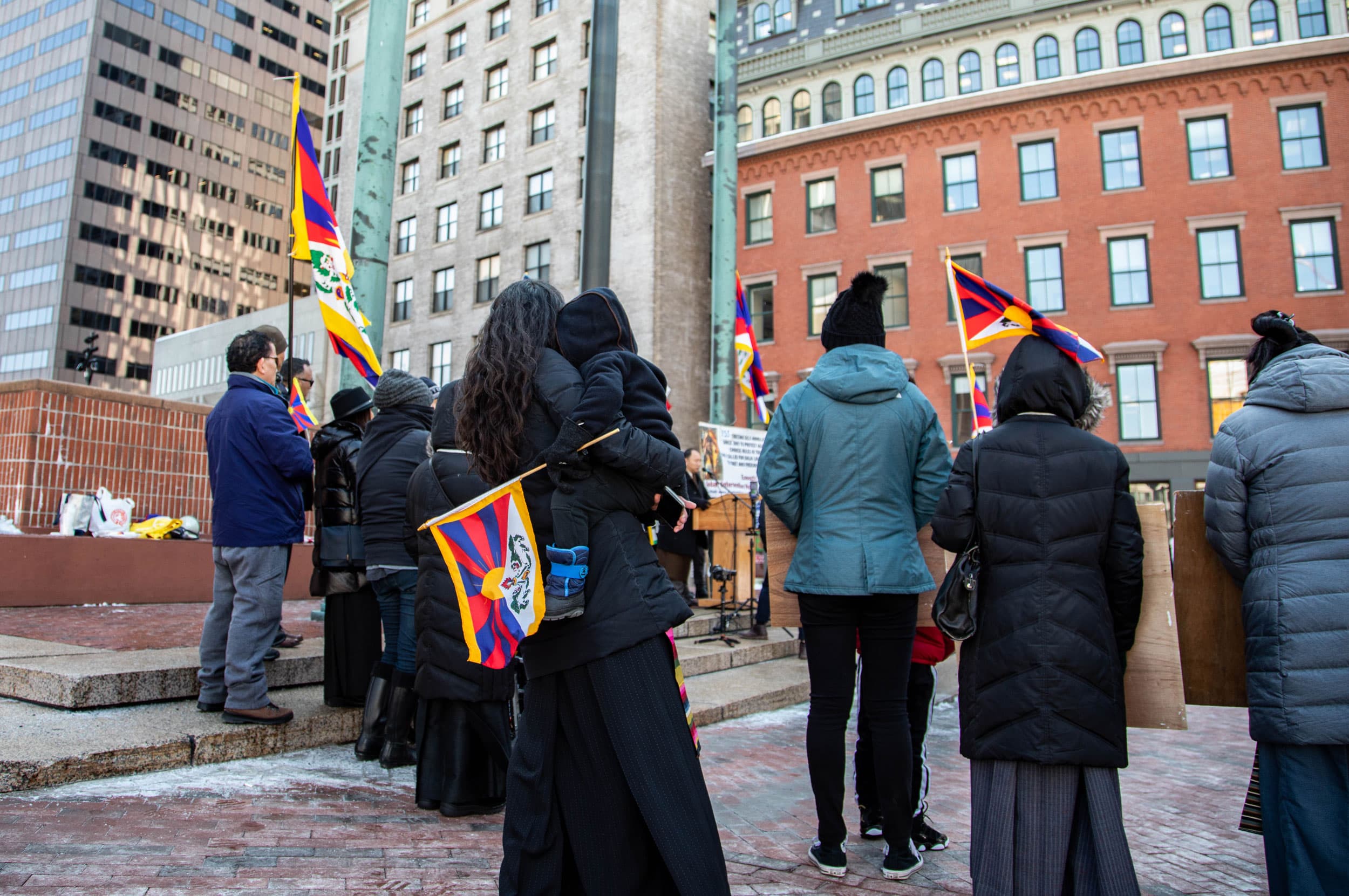 A woman stands with her back to the camera and holds a young child. A group of people is watching a flag move up a flagpole.