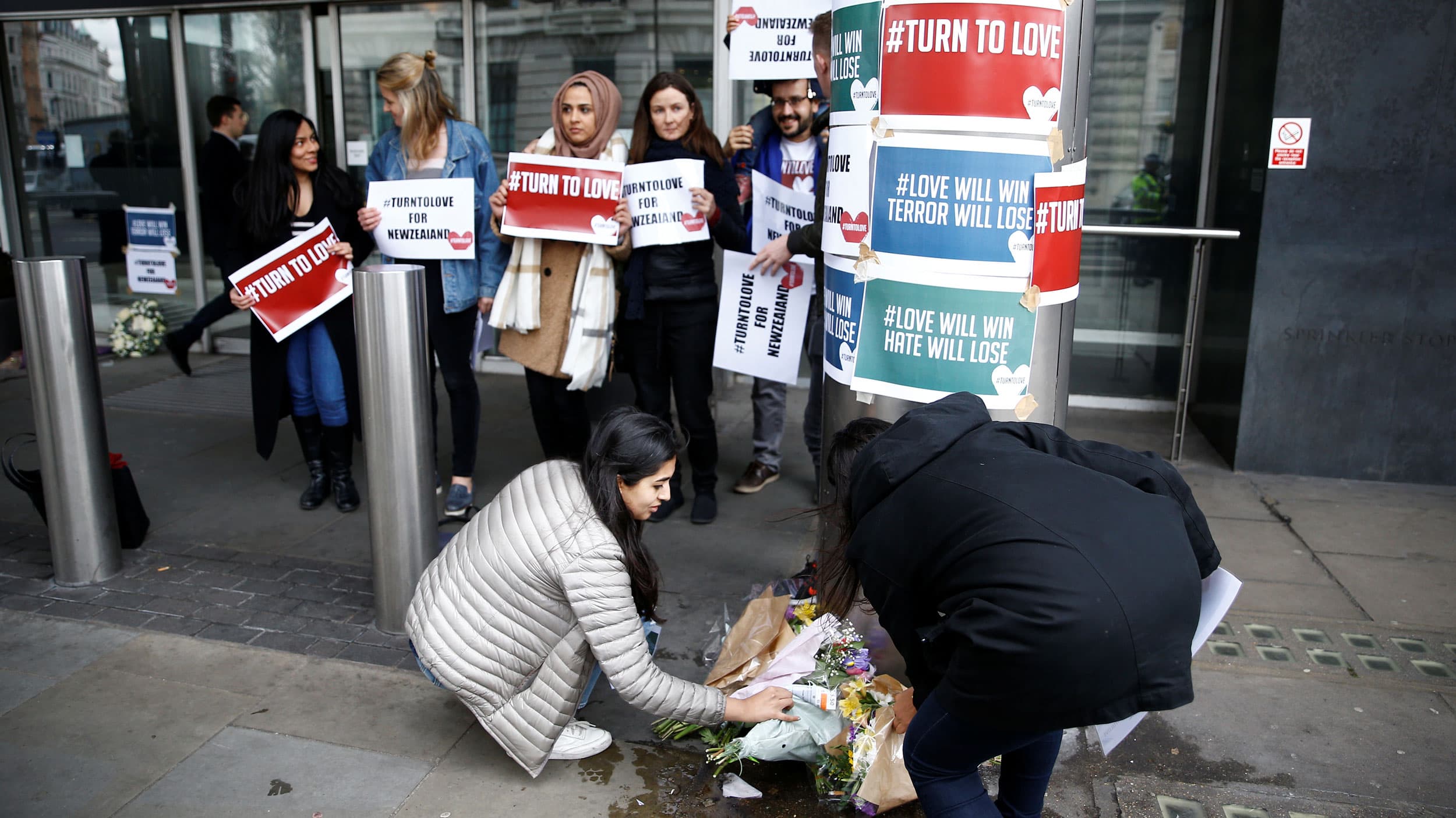 A group of people leave flowers outside a building with signs table to a column that say "#turntolove" and "#love will win terror will lose"
