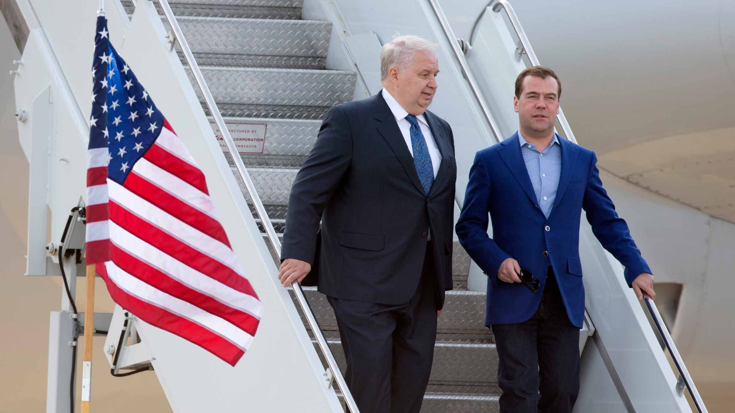 A man with white hair walks down the steps to a small plane with another younger man,