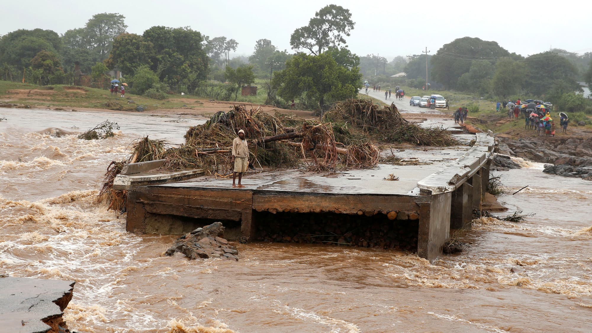A man is shown standing with a hooded jacket looks at a washed away bridge.