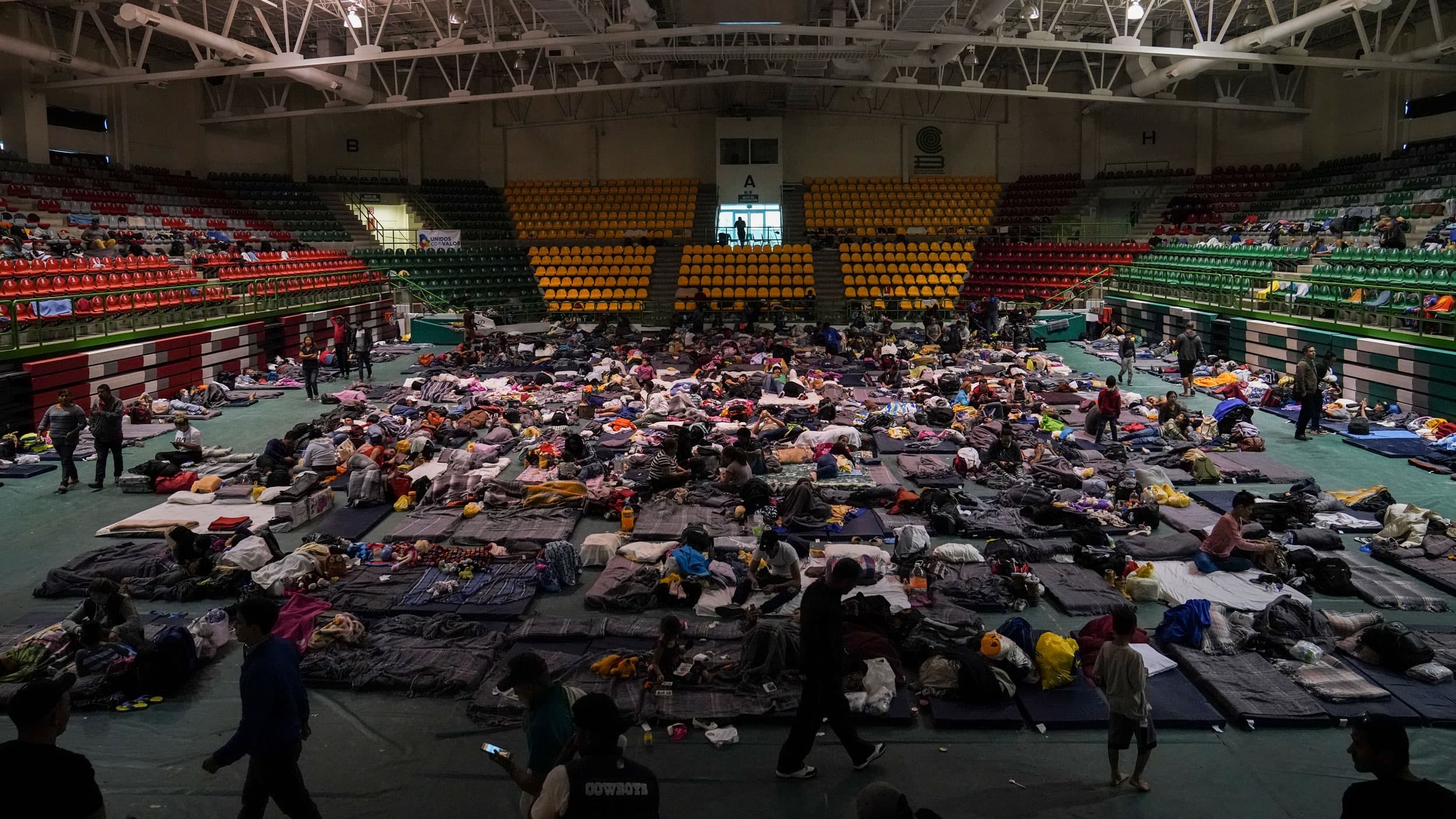 Migrants sitting and laying on mats are shown spread across a high school gymnasium floor.