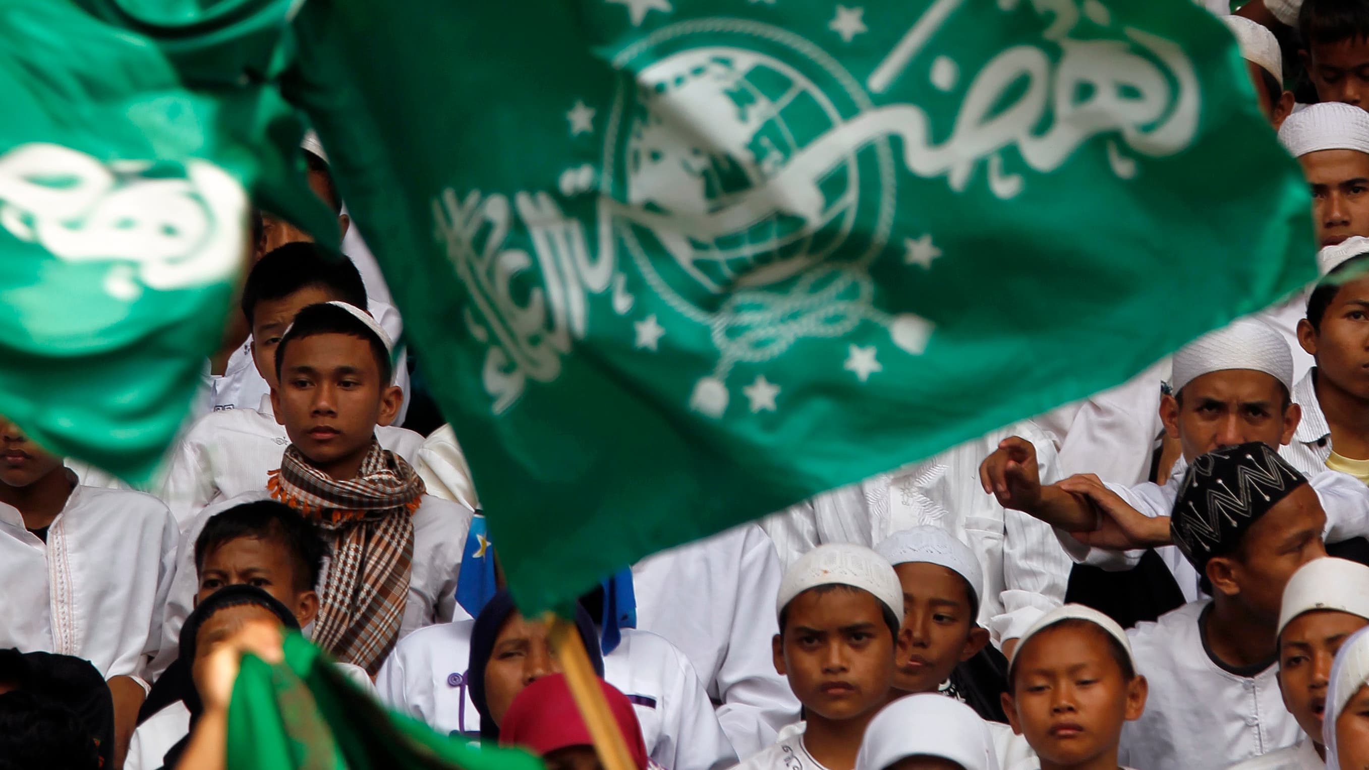 Young boys wearing white robes and caps stand behind green flag with Arabic script.