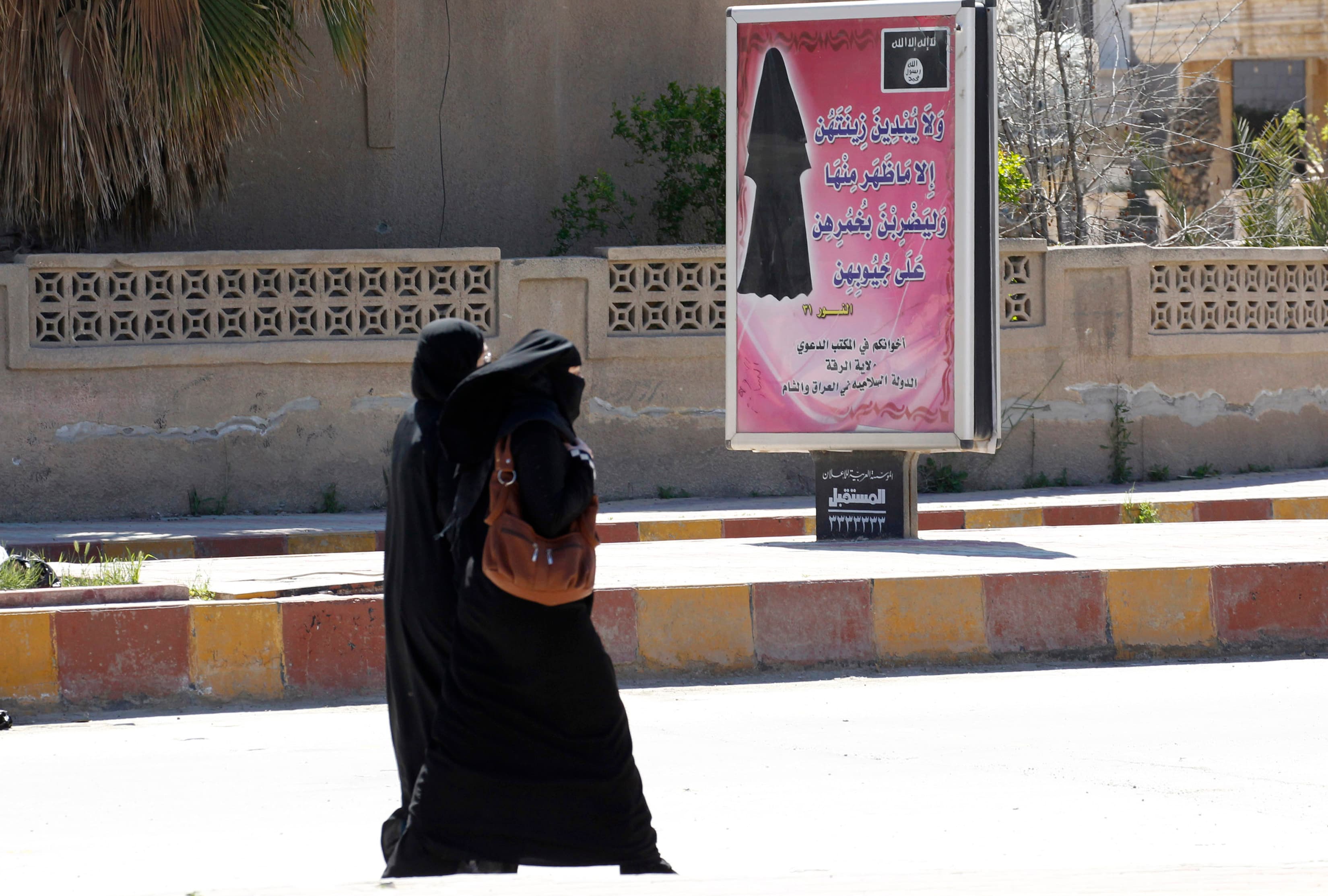 Two women wearing black veils walk outside past a sign with Arabic text.