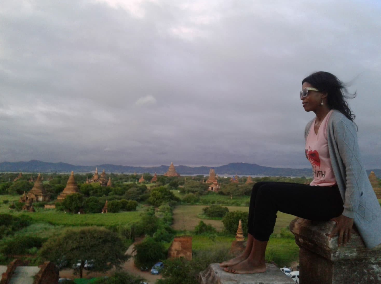 A woman sits atop a stone temple. Behind her are the tops of more temples in the distance.