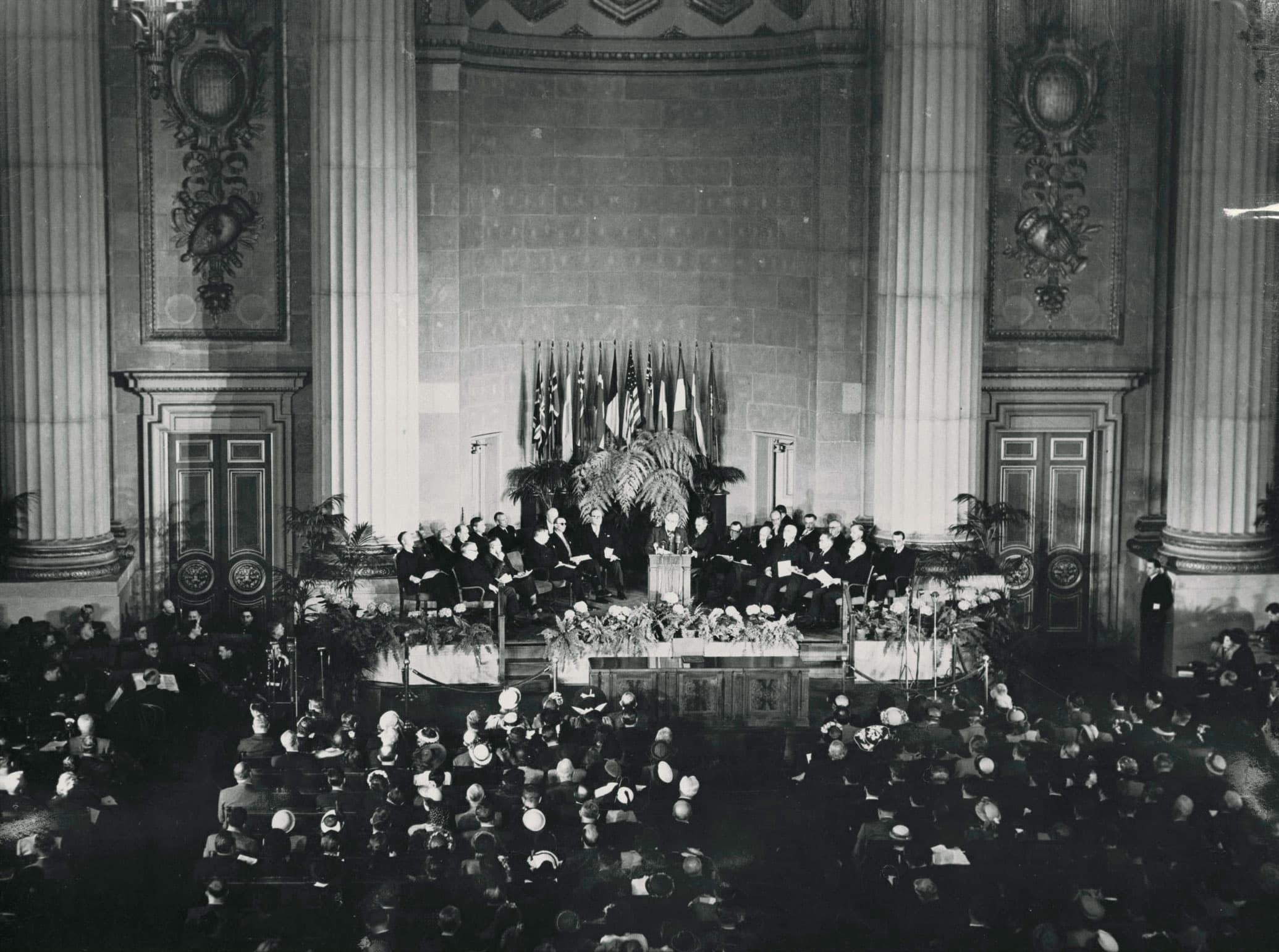 A historic photos shows dozens of delegates assembled in chairs around a podium, with more people seated around a lecturn. Behind there are giant columns.