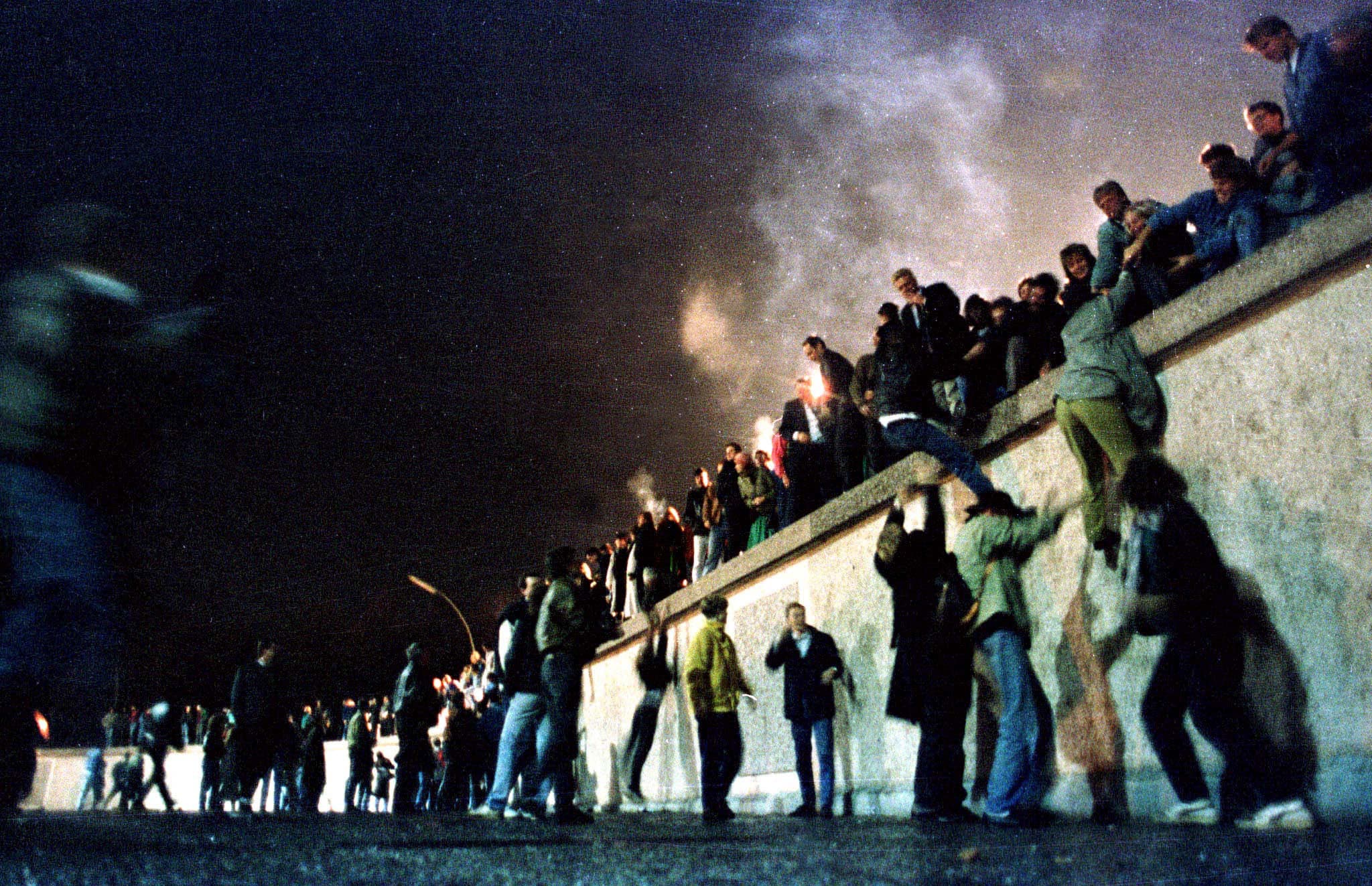 A crowd of people stand atop the Berlin Wall near Brandenburg gate in this 1989 photo. Behind them is smoke from what could be burning fires.