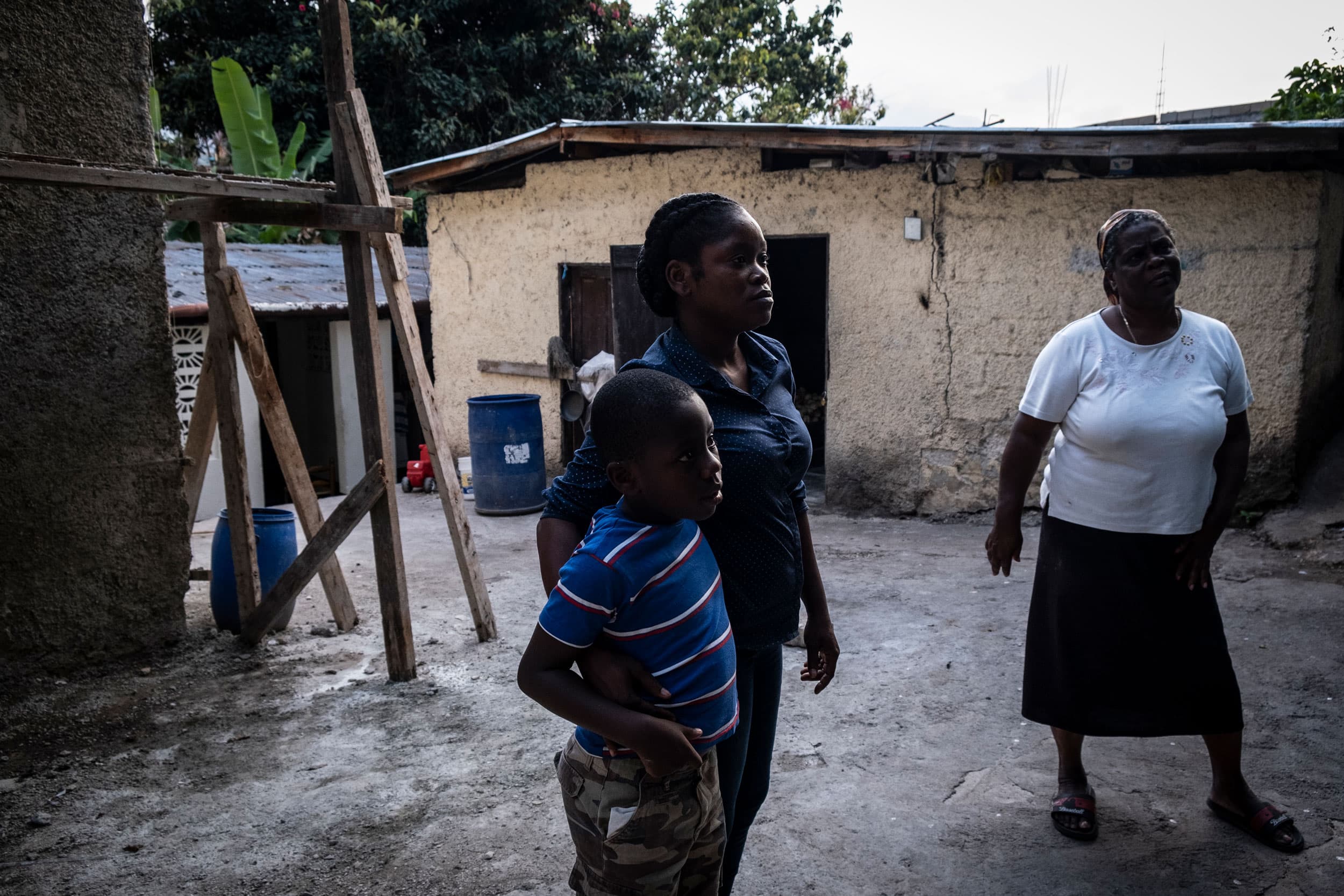 Three people stand in a courtyard. Behind them are simple buildings held up with blocks of wood.