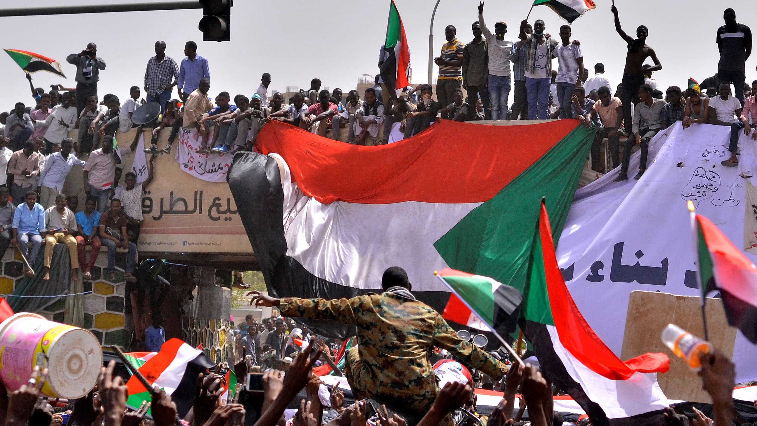 A thick crowd of people wave Sudanese flags and celebrate. A man in military fatigues is being carried through the crowd.