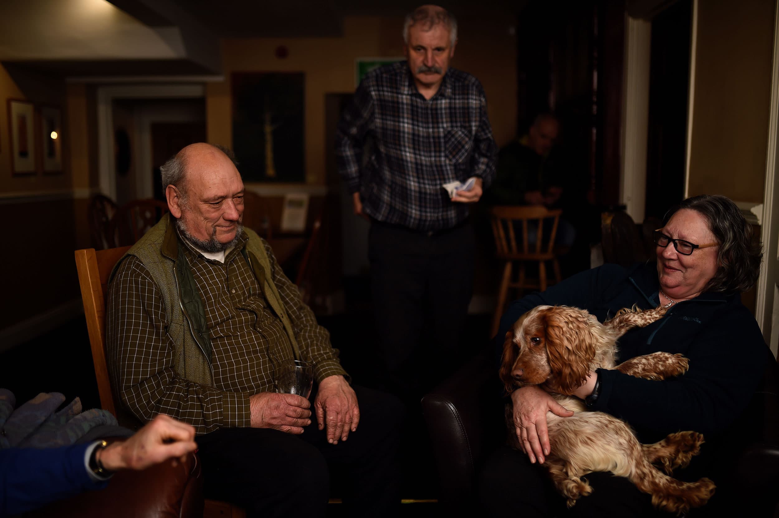 An older man is shown sitting down, wearing a vest and holding a glass next to a woman holding a dog.