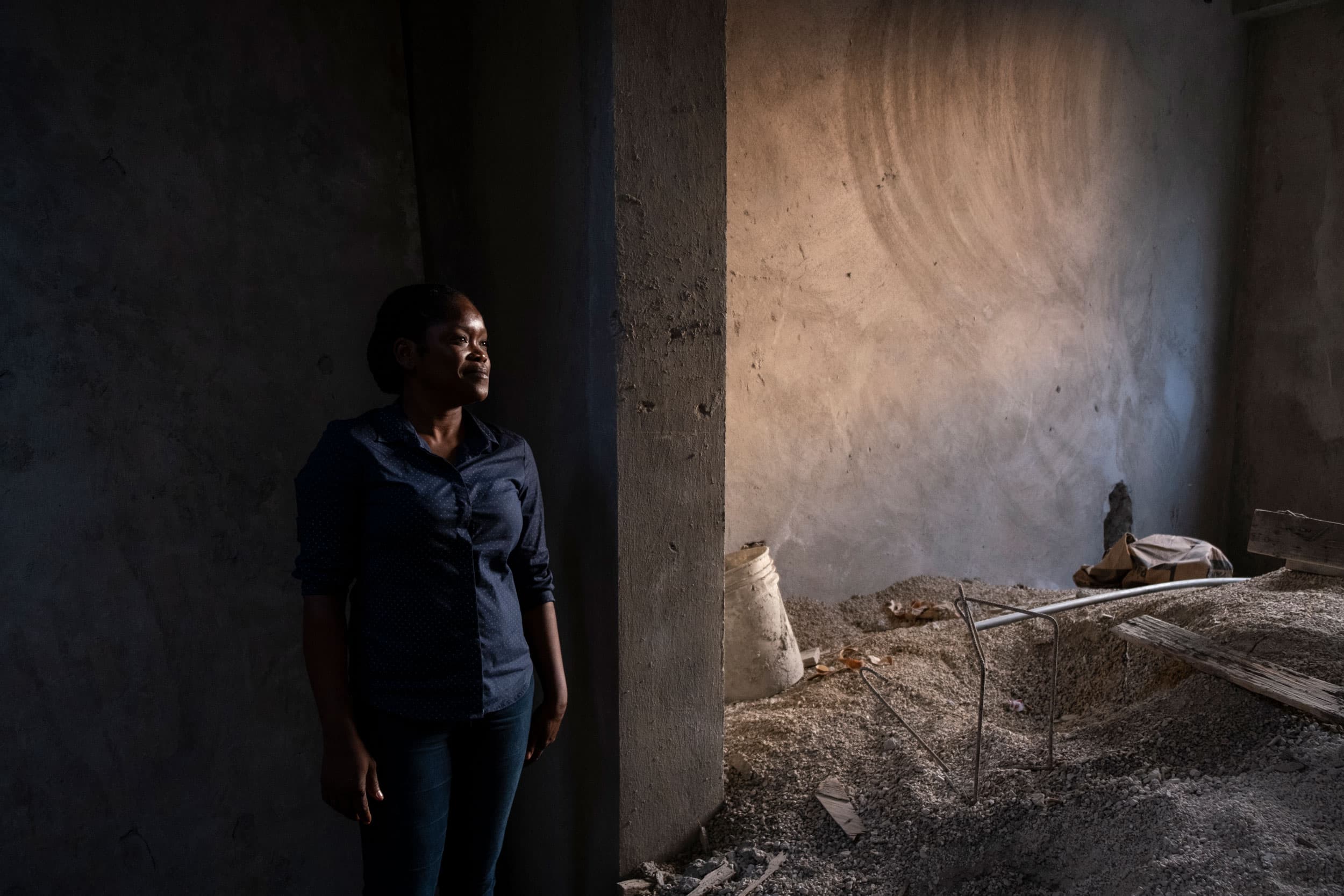 A woman is shown standing among the construction materials of her home with cement walls.