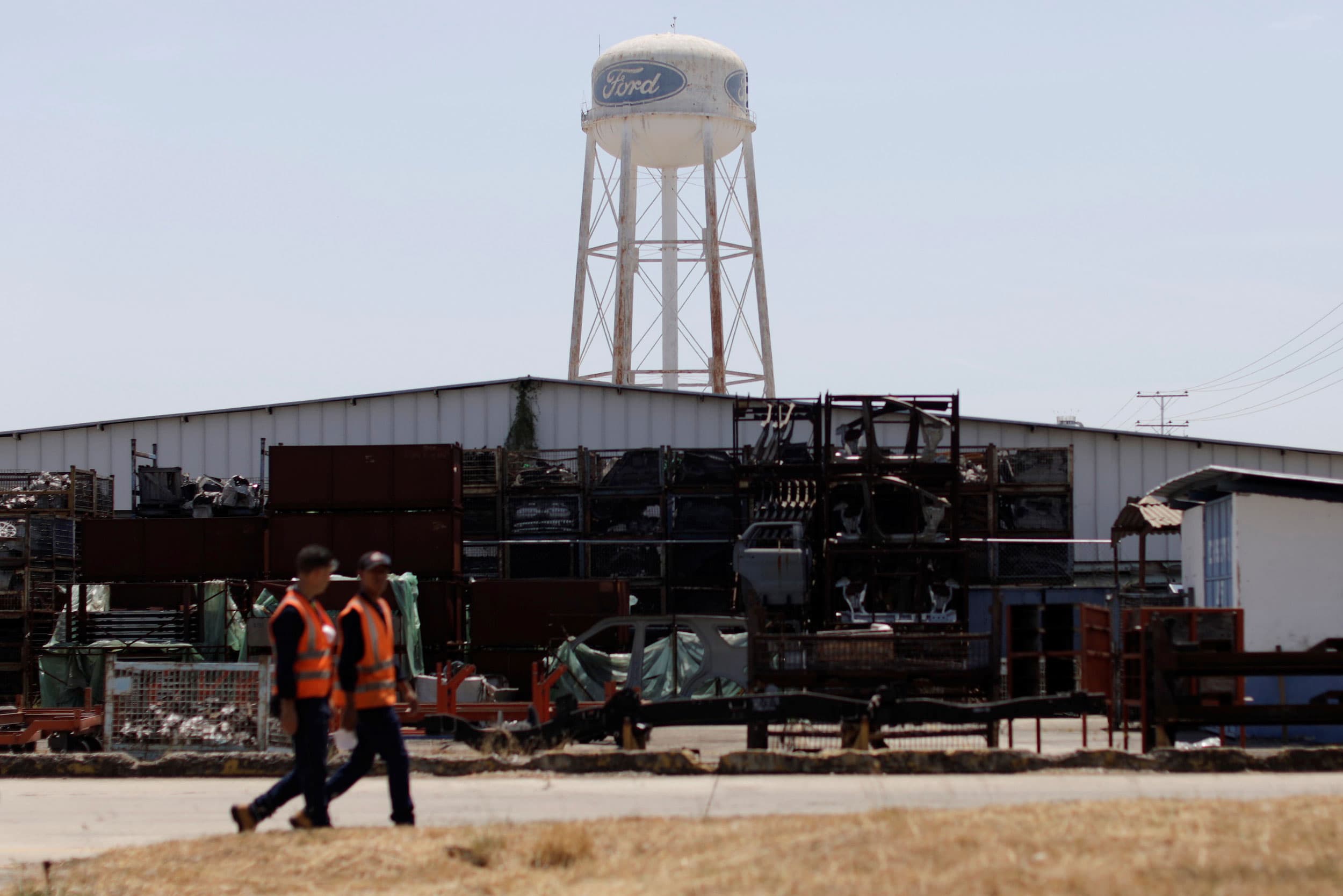 Two men are shown wearing reflective safety vests while walking at a Ford plant.