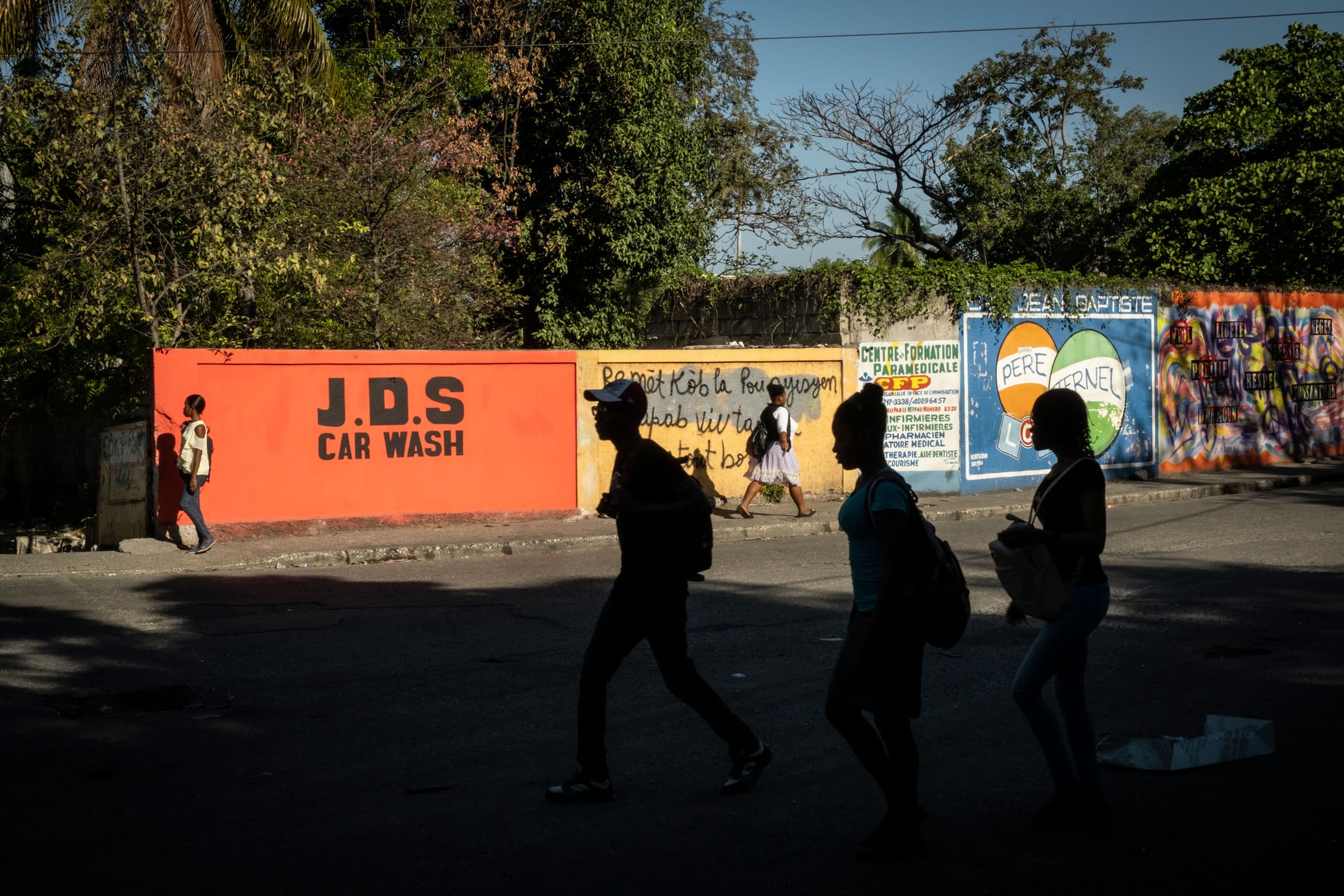 Several people are show in shadown walking past a wall in full light and painted with graffiti.