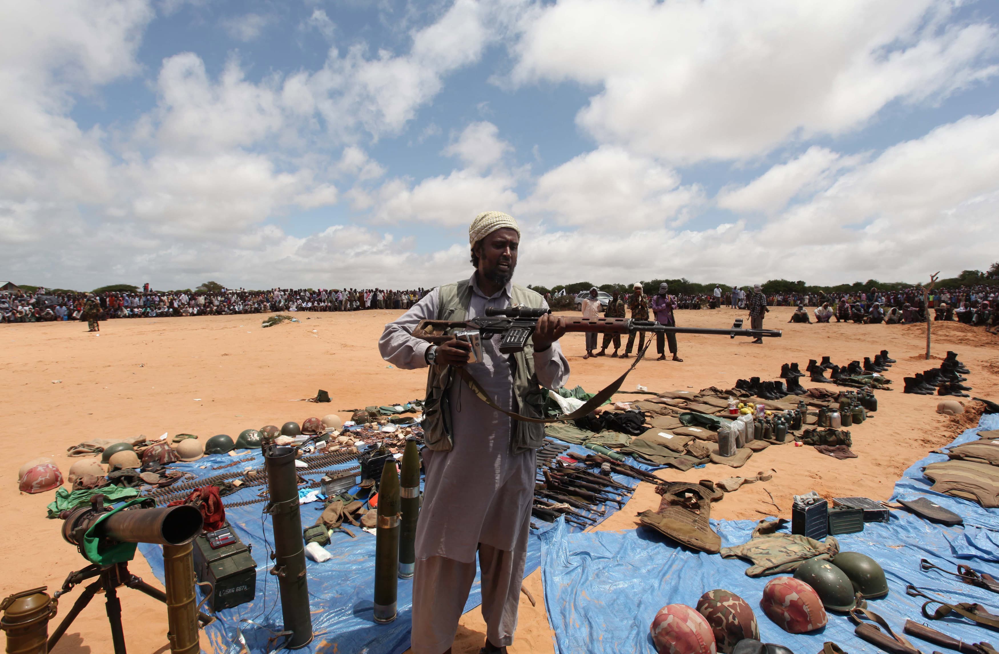 Man stands near weapons laid out on tarps in the desert.