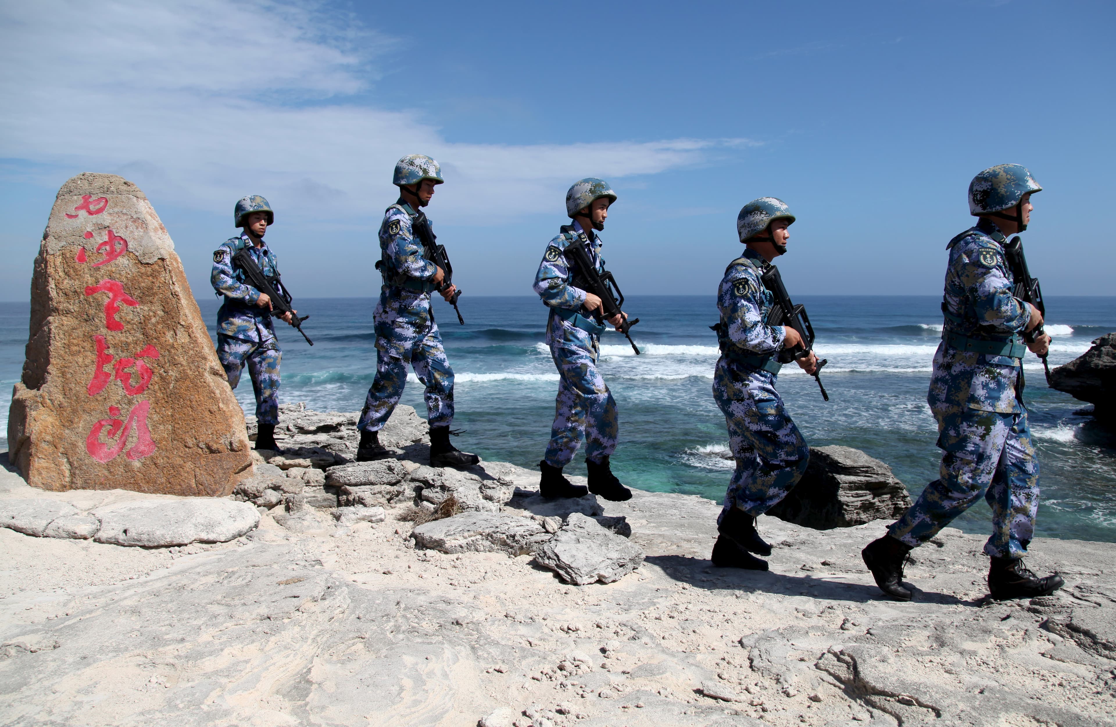 Chinese soldiers march in single file wearing blue and grey uniforms near a rock with red Chinese letters.