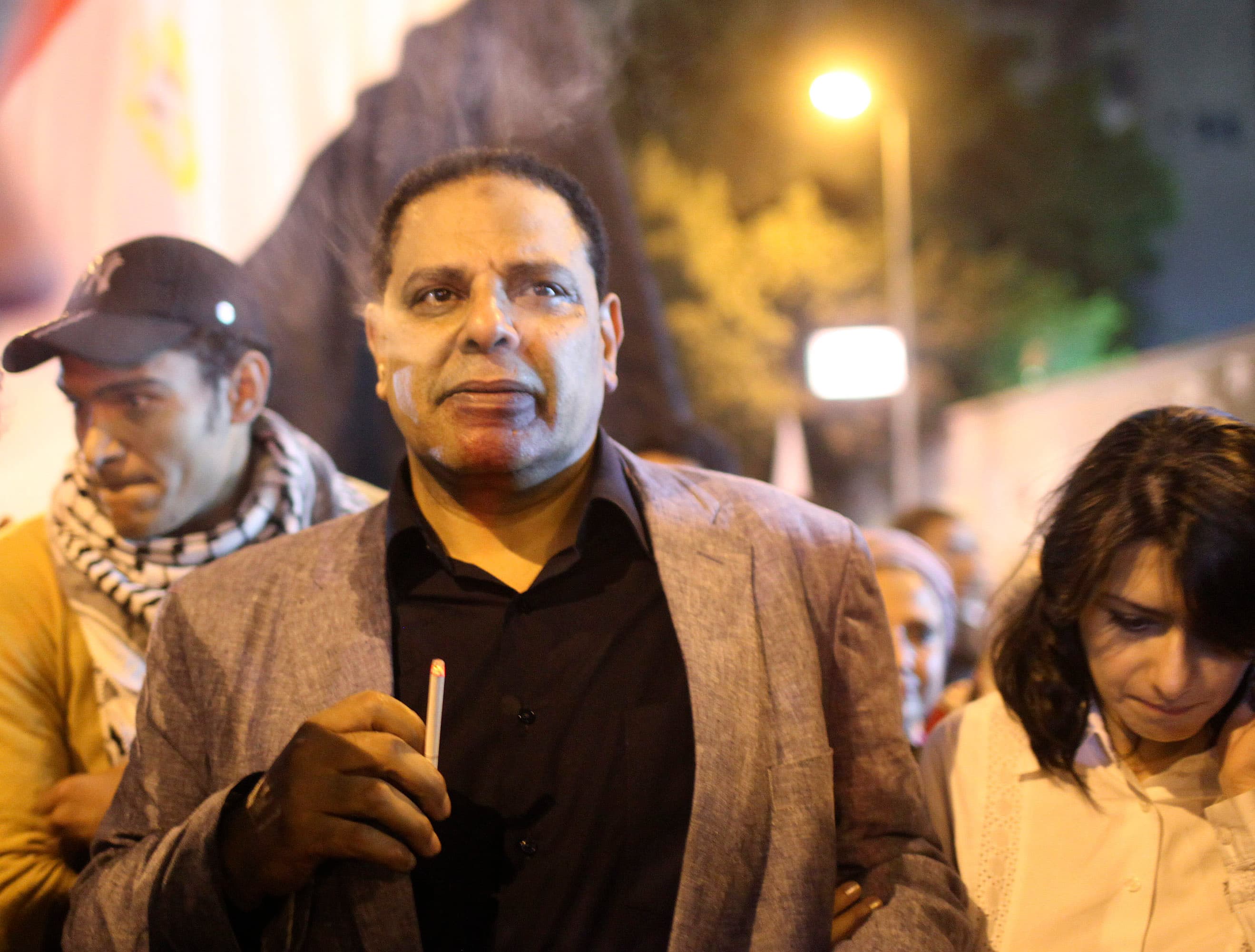 A man walks with a crowd at night with the Egyptian flag behind him.