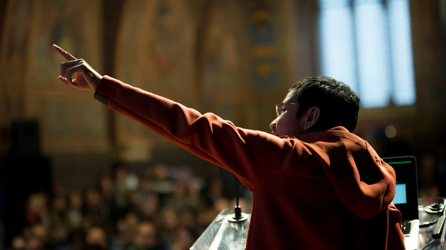 A woman in an orange hoodie has her back to the camera and is pointing to the left. Behind is a blurred collection of faces.