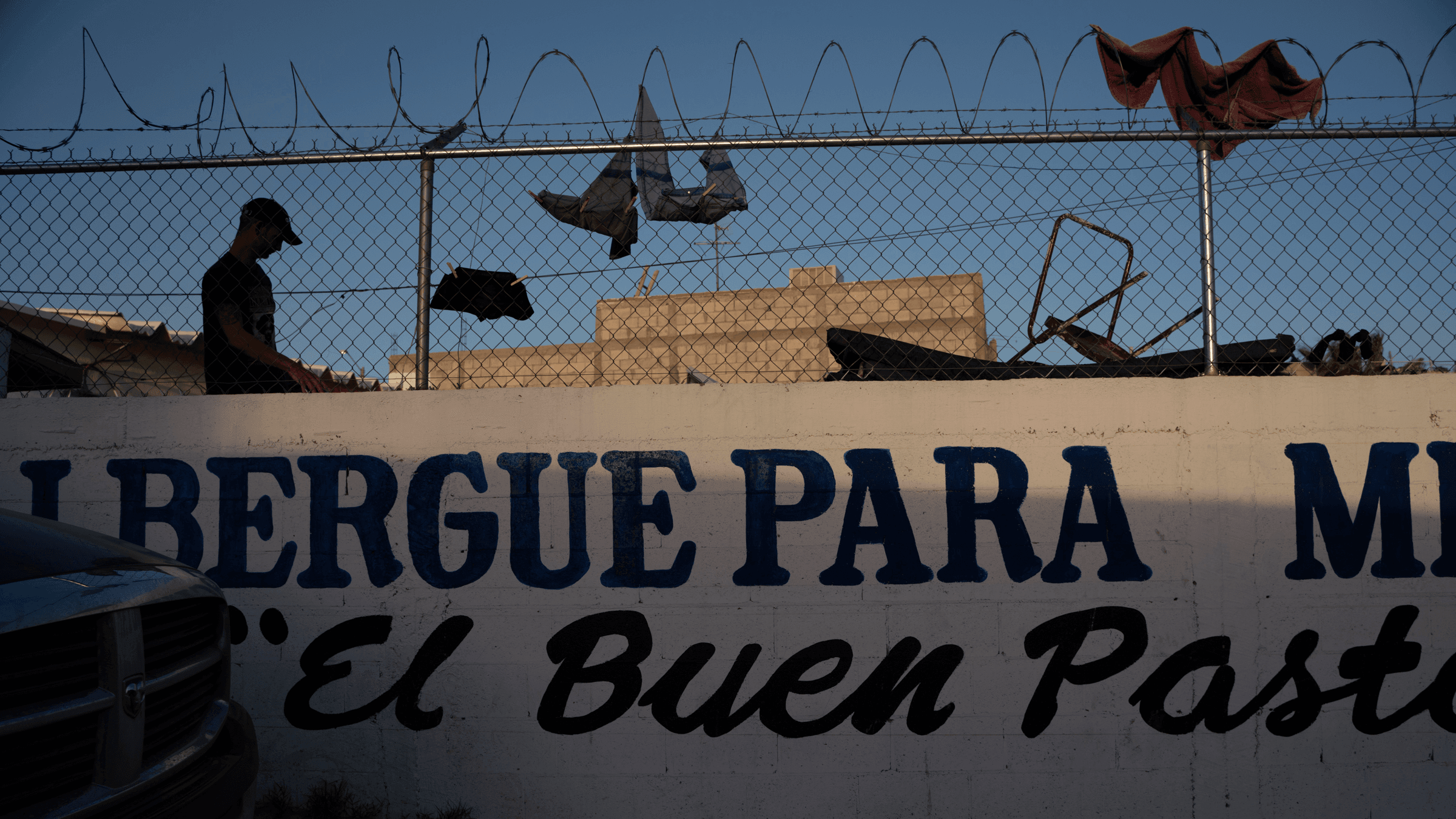 A man is silhouetted against a fence topped with razor wire. People have clothespinned clothing on the fence to dry.