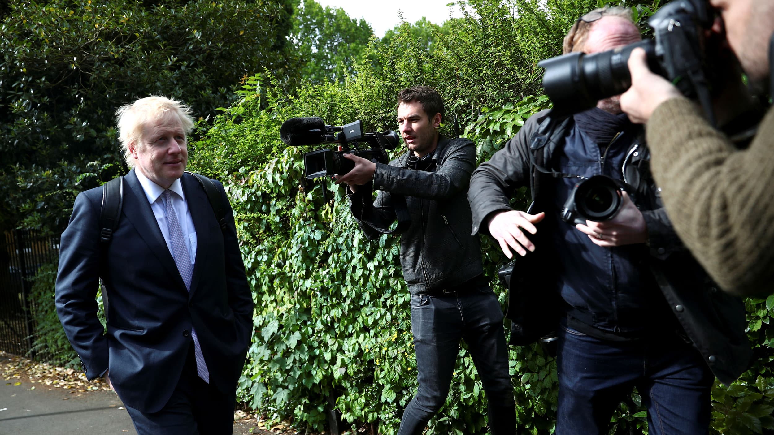 Boris Johnson is shown walking with his hands in his pockets and a gaggle of photographers around him.