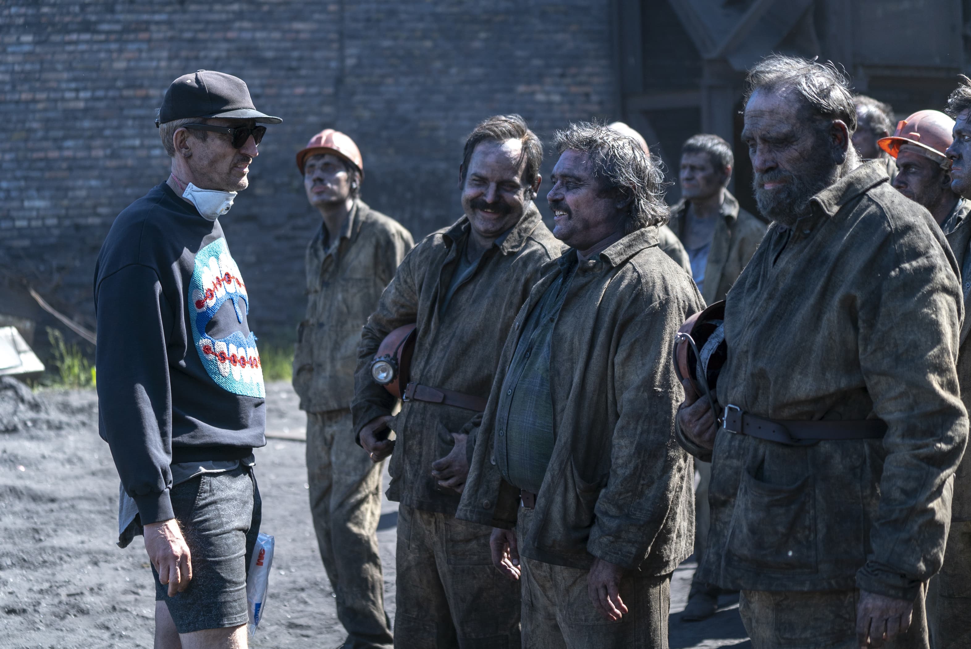 A man in shorts and a baseball hat stands next to construction workers covered in soot.