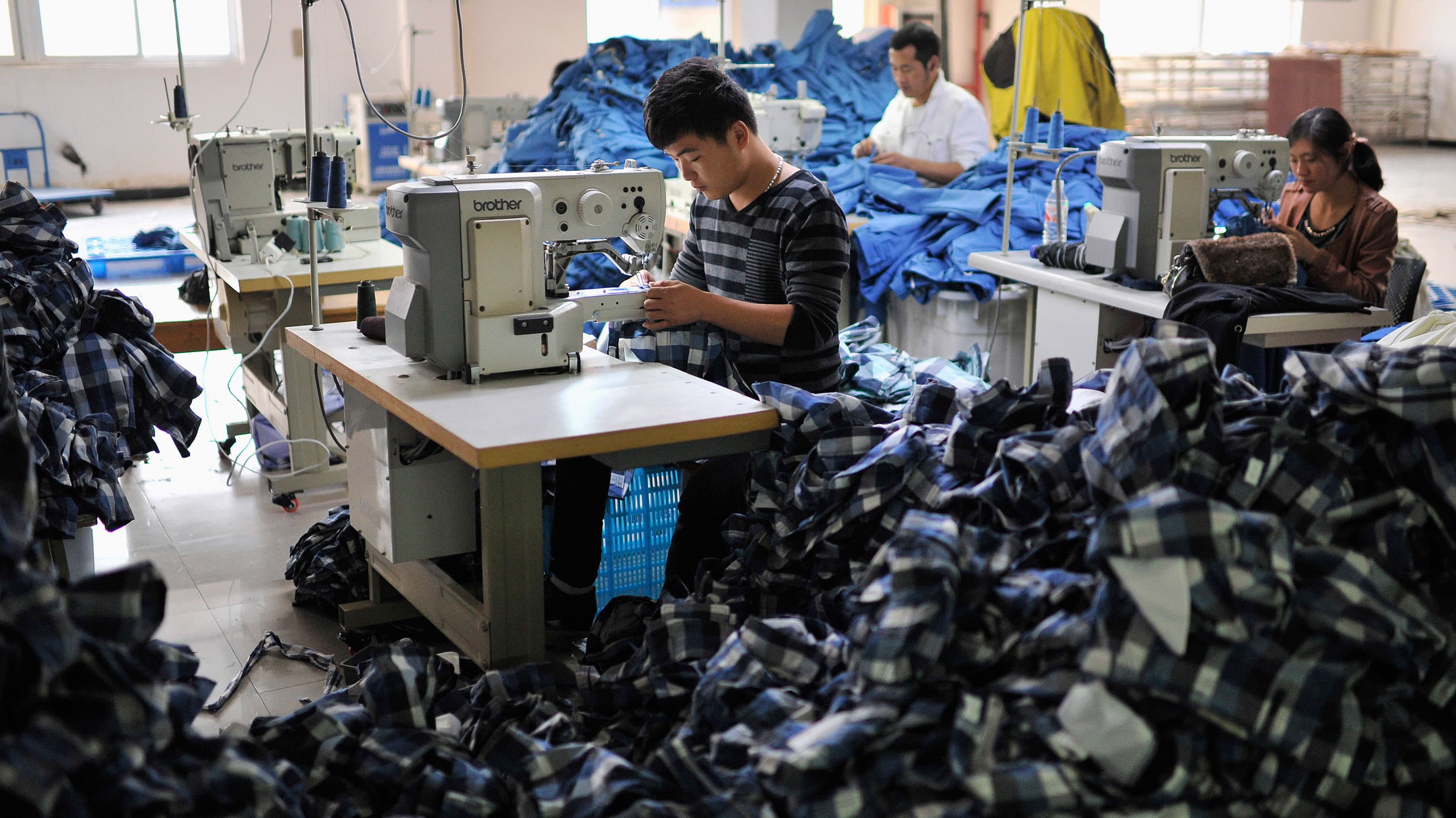 A man sits at a sewing machine table and sews a blue plaid button up shirt. In front of him sits a large pile of identical shirts.