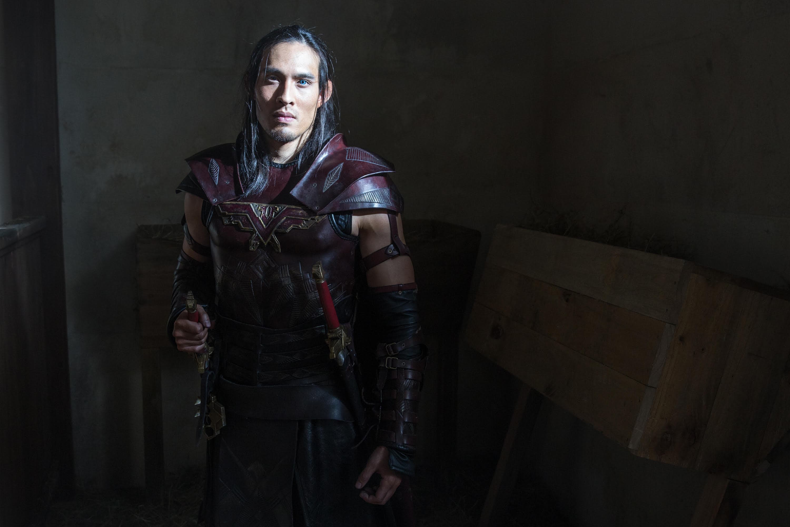 A man in an armor is looking straight at a camera. He's holding a sword