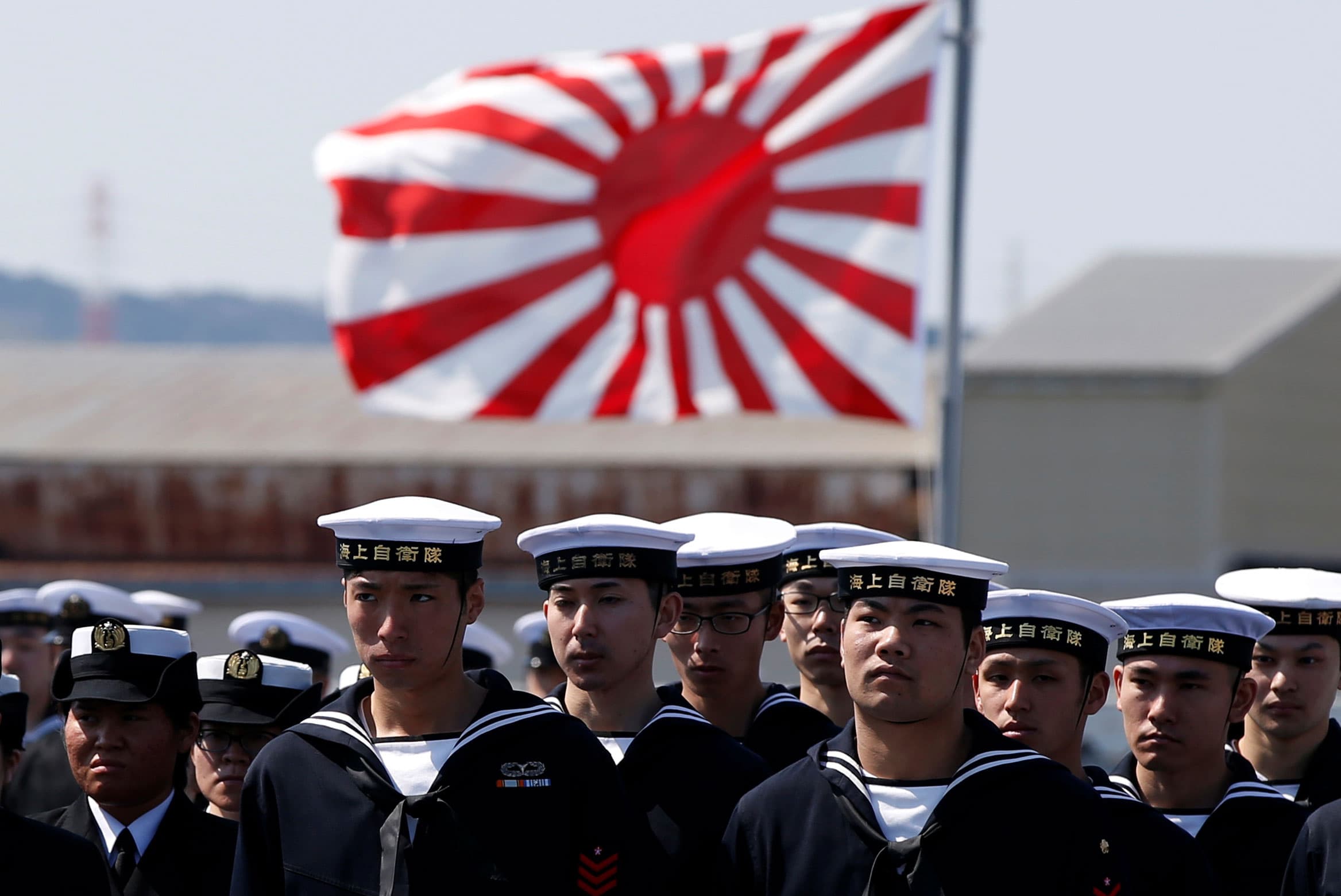 Sailers stand in formation and wear white hats. Behind them is a large flag.
