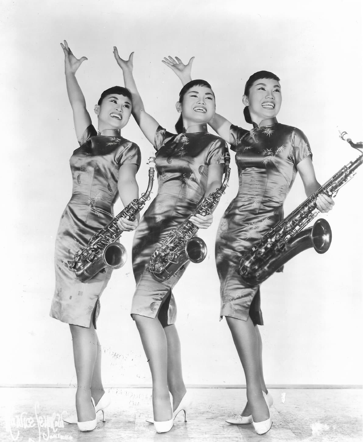 A black and white photo of three young Korean women posing with their hands up and holding saxophones.