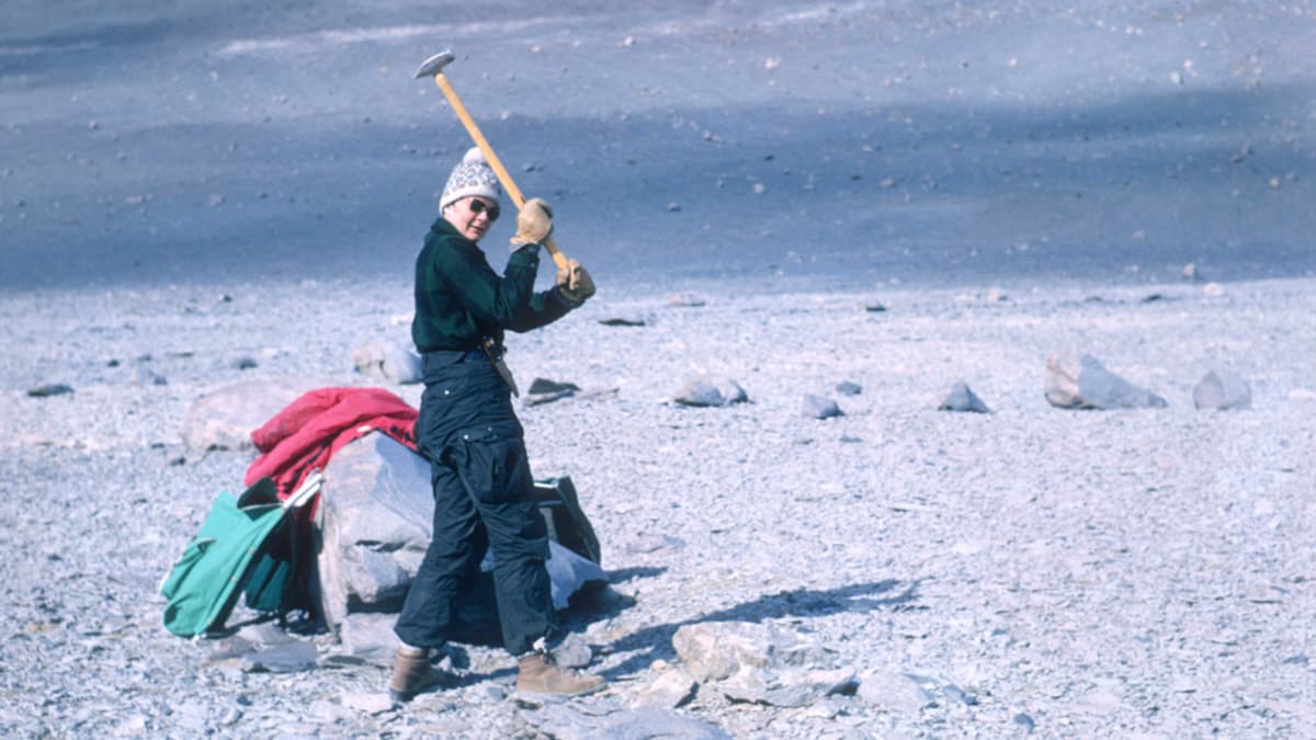 Woman wearing overalls and sunglasses holds and ax with both hands while looking at the camera.