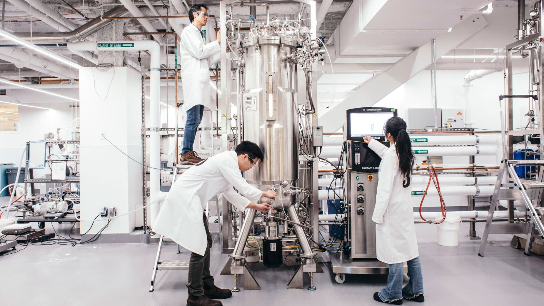 Three scientists in white lab coats stand around a tall metal container while taking measurements.